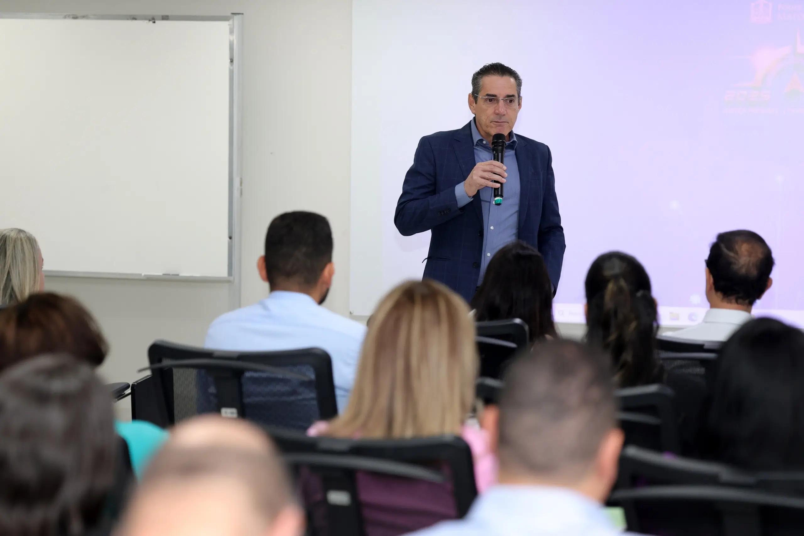 Homem de terno azul escuro e camisa azul fala ao microfone diante de participantes sentados em uma sala, com projeção ao fundo.