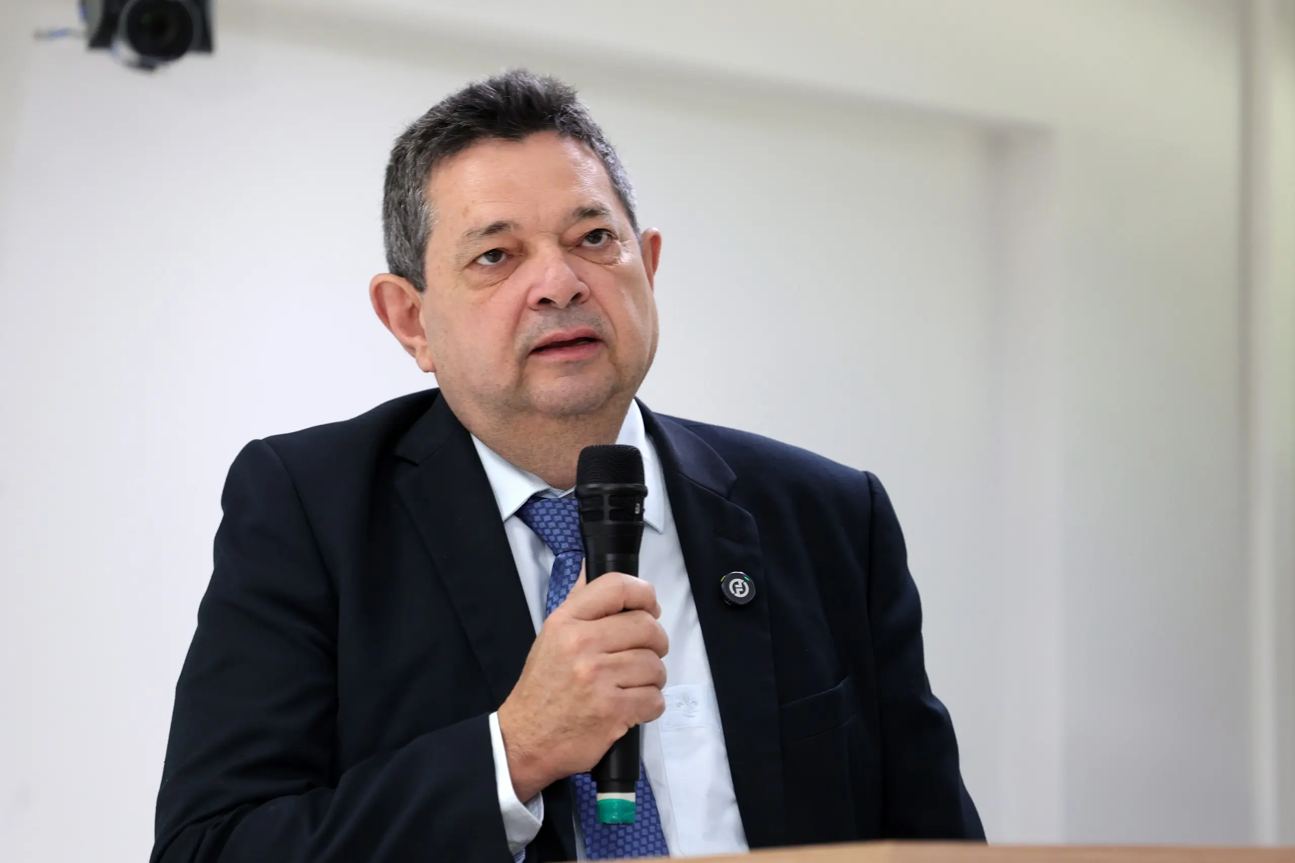 Homem de terno escuro, camisa branca e gravata azul fala ao microfone durante evento realizado em ambiente interno.