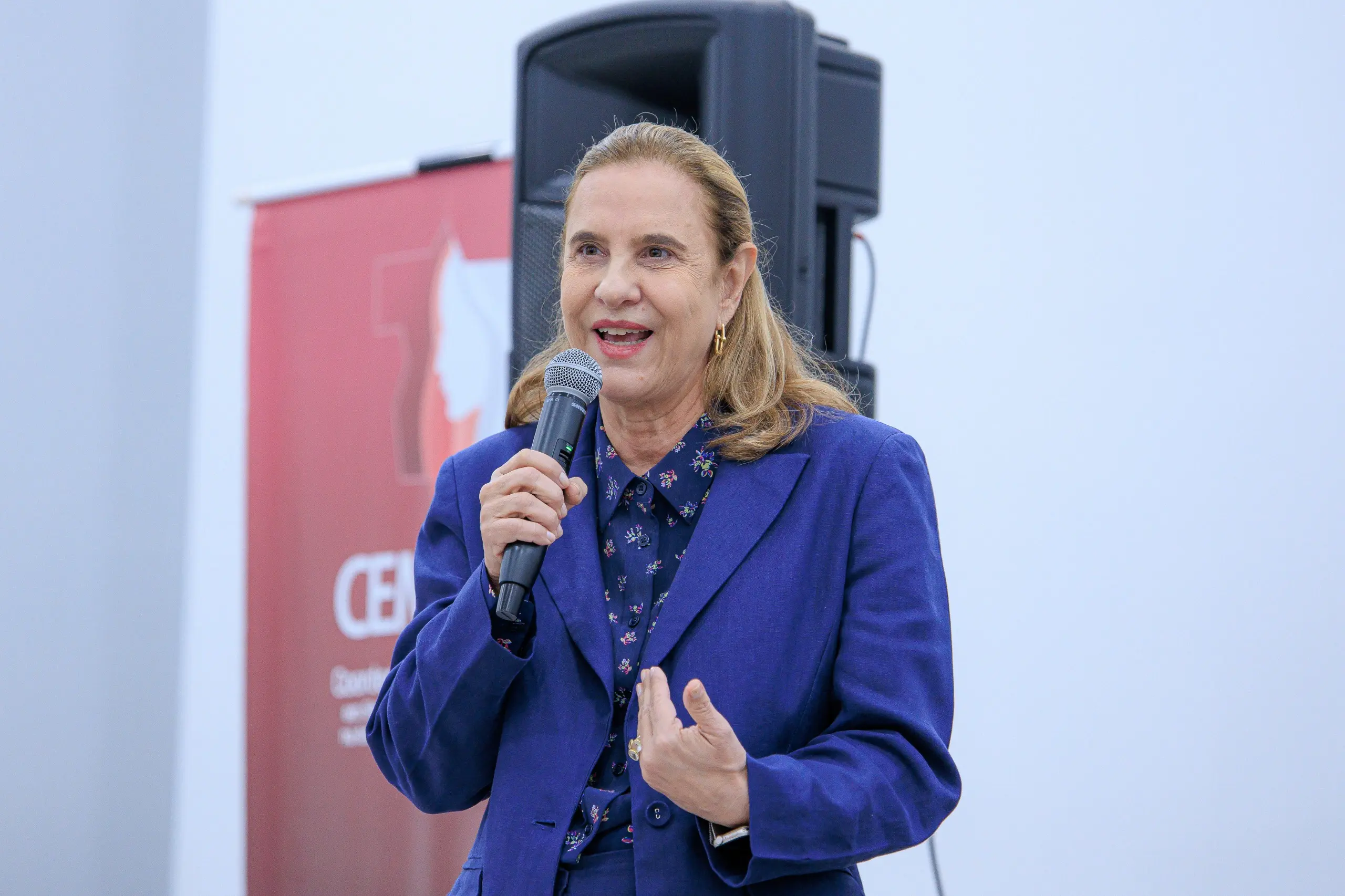 Mulher de cabelos loiros, blazer azul e camisa estampada, fala ao microfone e gesticula com a mão. Ao fundo, banner vermelho do CEMULHER e caixa de som