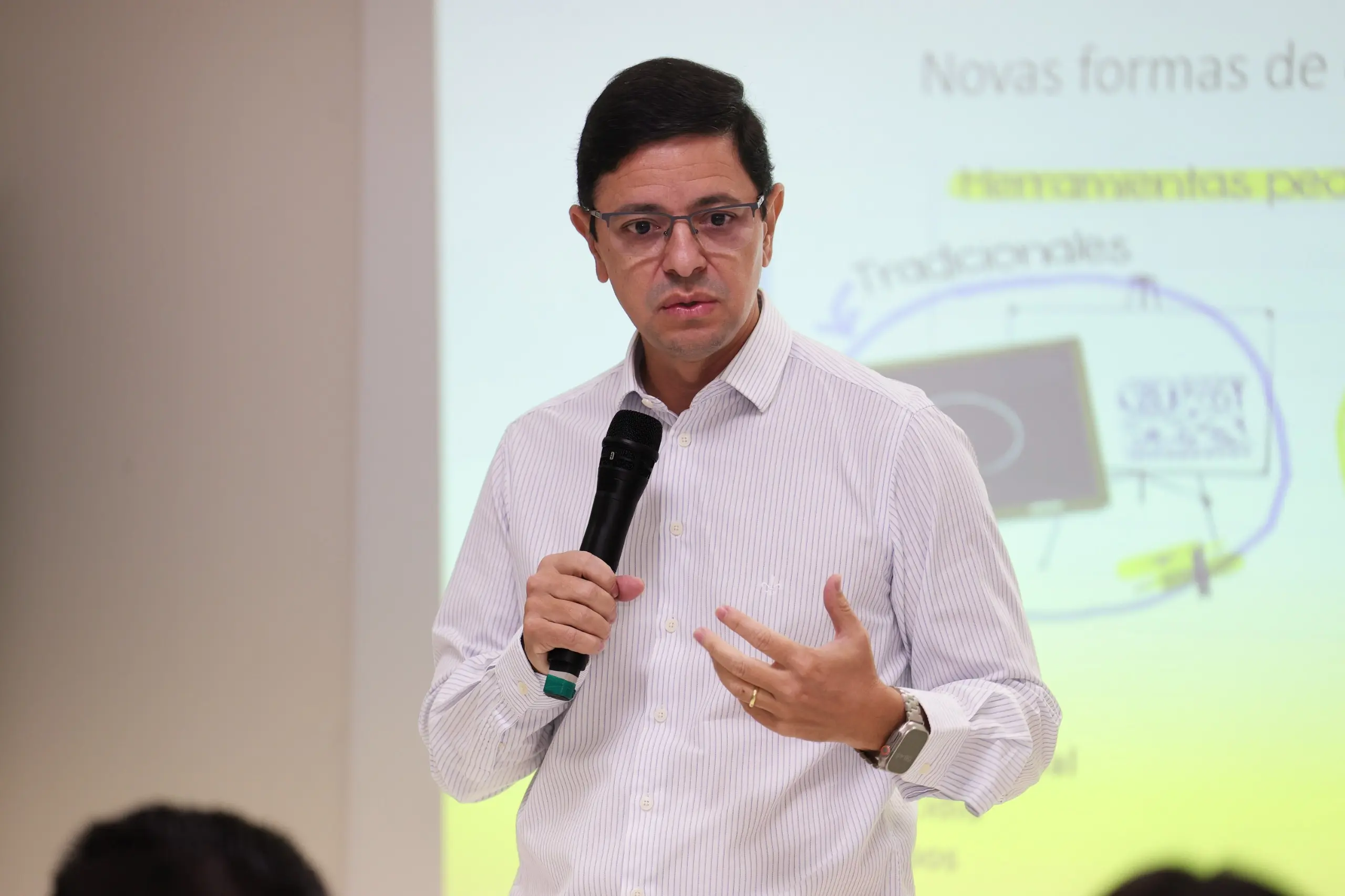 Homem de camisa social clara e óculos fala ao microfone durante apresentação. Ao fundo, há uma projeção com texto e imagens destacadas em amare