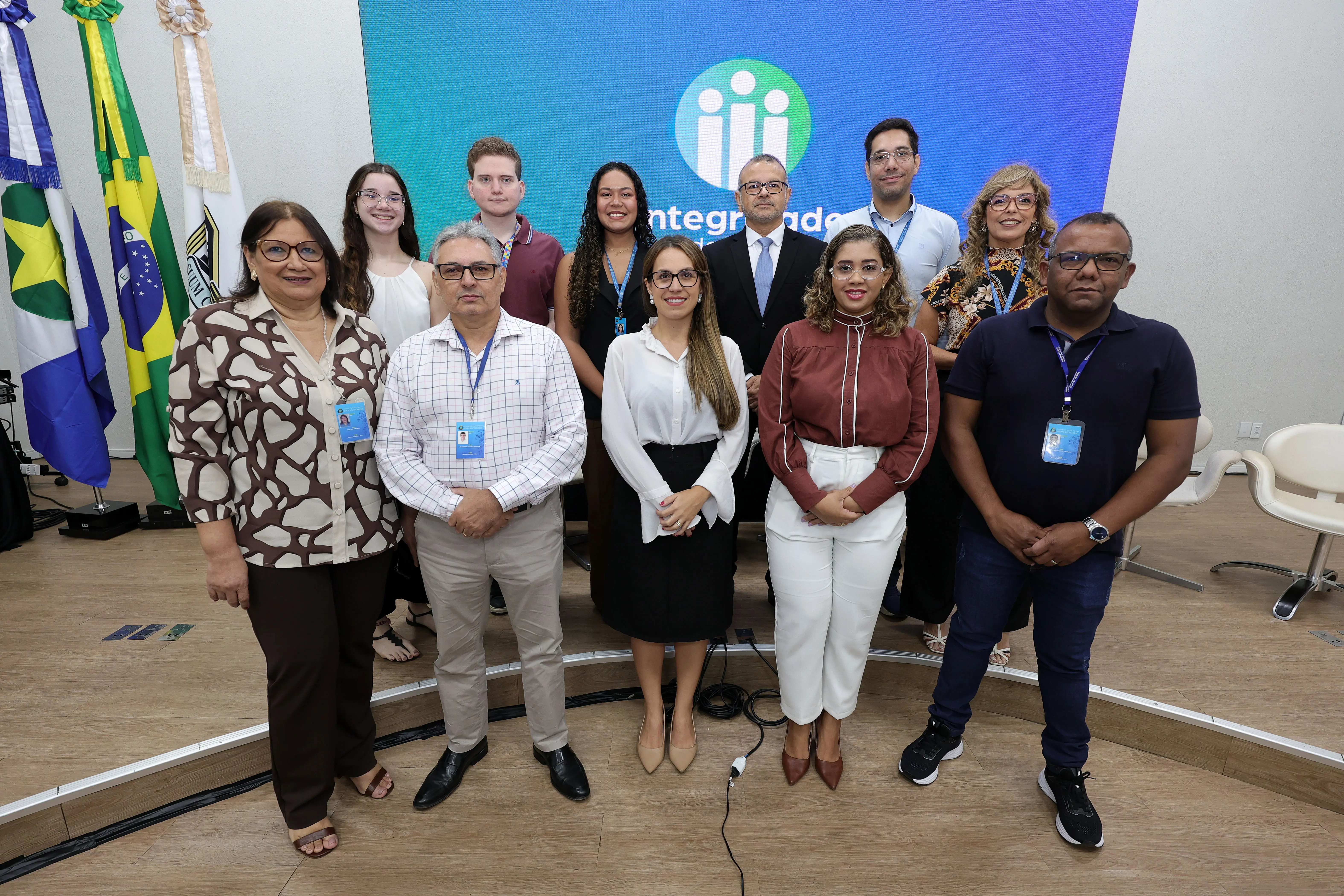 Grupo de homens e mulheres posa para foto no palco de um auditório, em frente a bandeiras oficiais e um telão azul. Usam trajes esporte fino e crachás de identificação no pescoço.