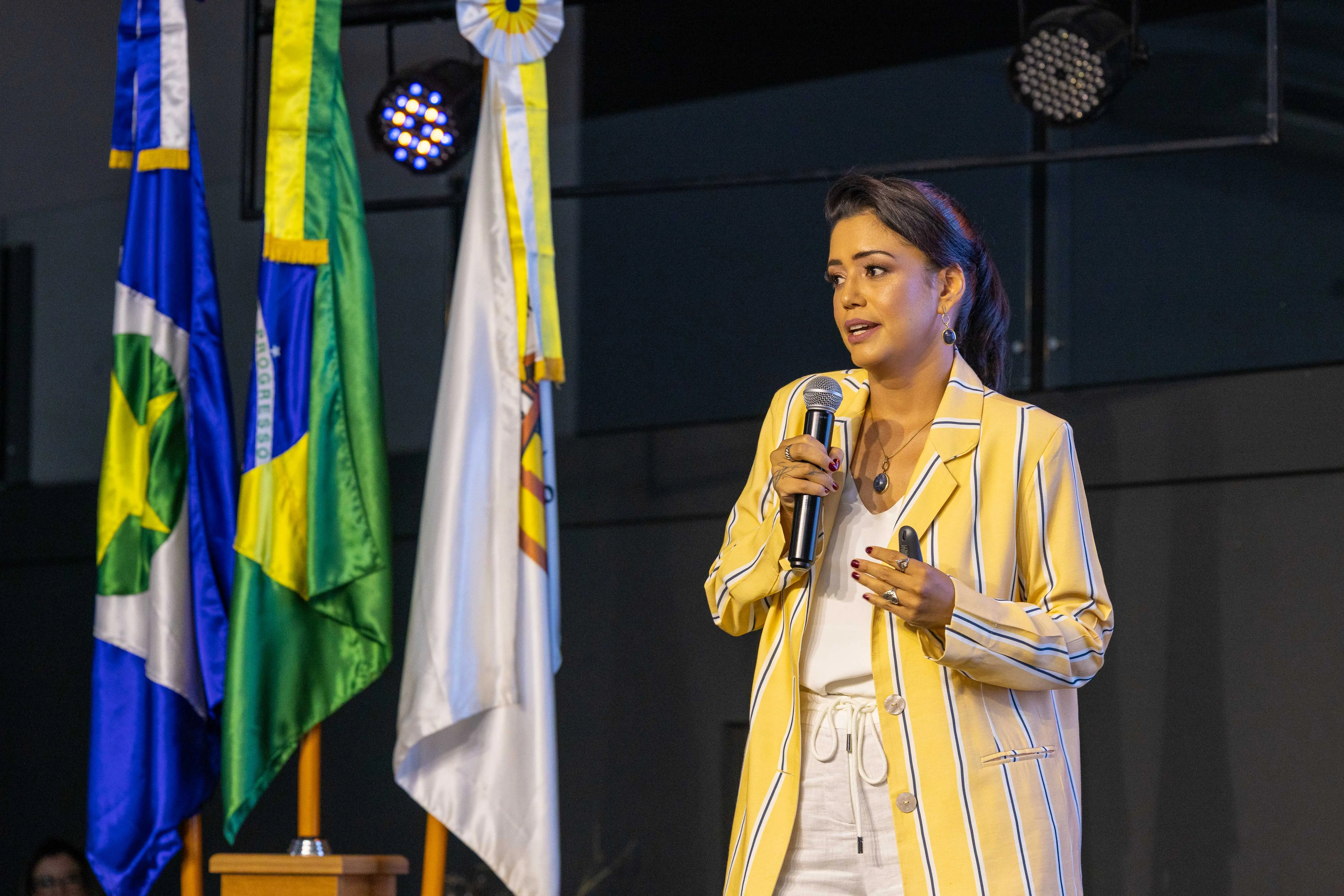 Mulher fala ao público durante o evento TJMT Inclusivo, em Rondonópolis. Ela segura um microfone e usa blazer amarelo com listras brancas, com bandeiras do Brasil e do Judiciário ao fundo no palco iluminado.