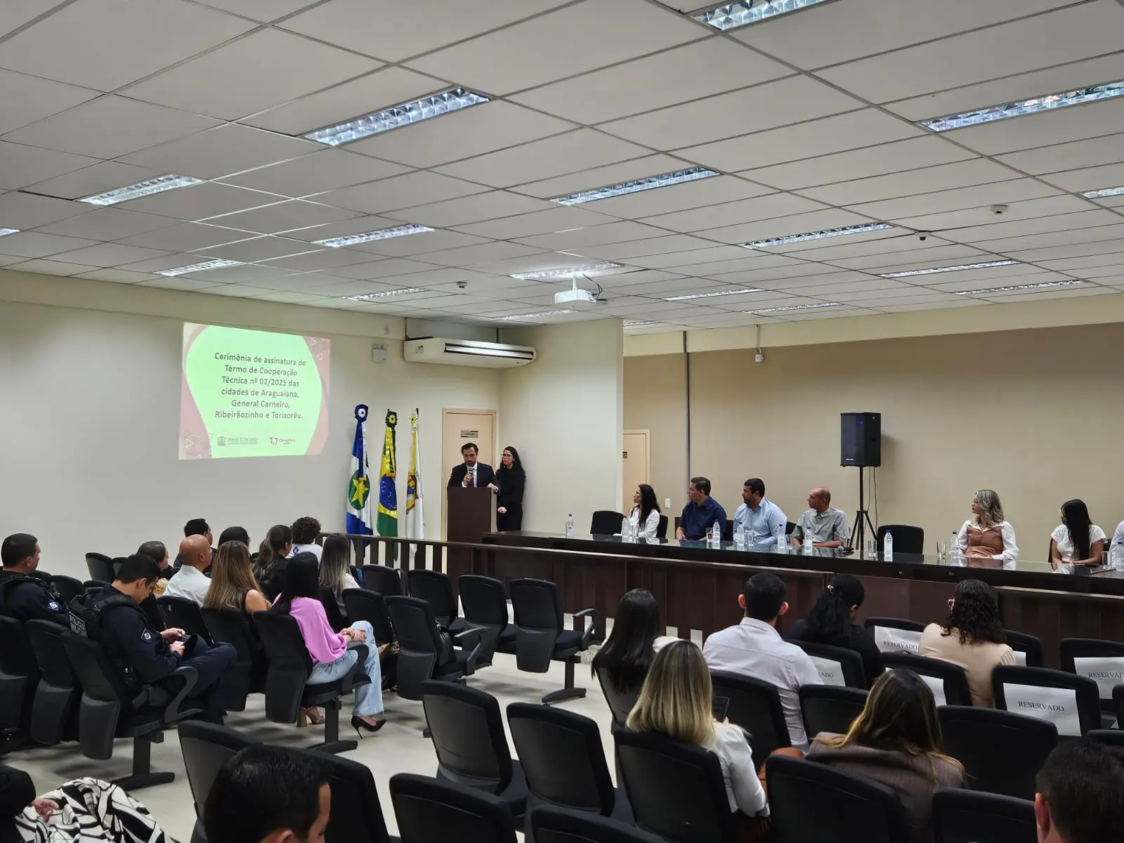 Auditório interno lotado com pessoas sentadas assistindo a uma cerimônia. Na frente, uma mesa com autoridades. Um projetor exibe "Cerimônia de assinatura do Termo de Cooperação Técnica...".