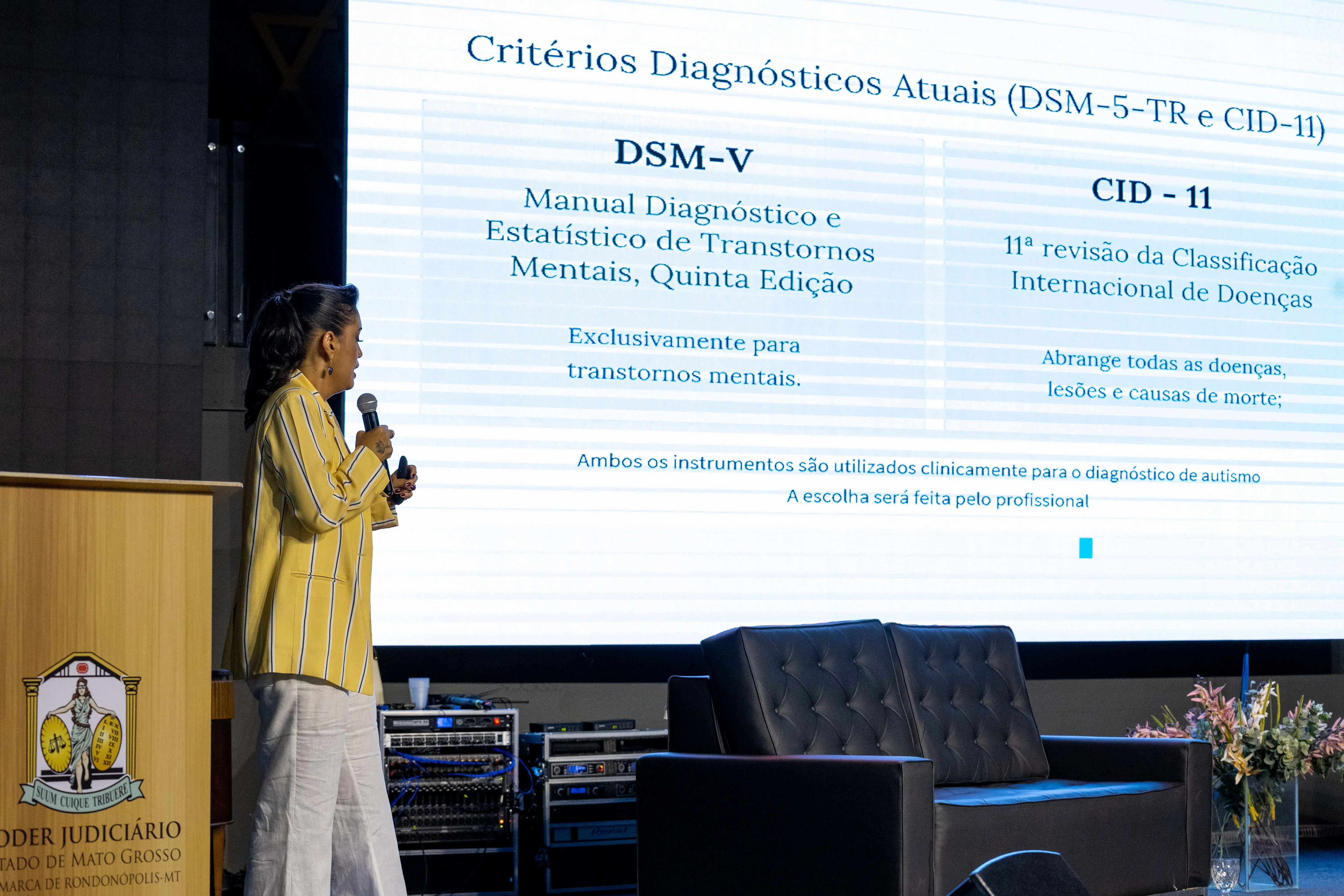 Mulher apresenta no palco do TJMT Inclusivo, ao lado do púlpito do Poder Judiciário de Mato Grosso. Na tela, aparecem definições dos manuais DSM-V e CID-11 usados para diagnóstico do autismo.