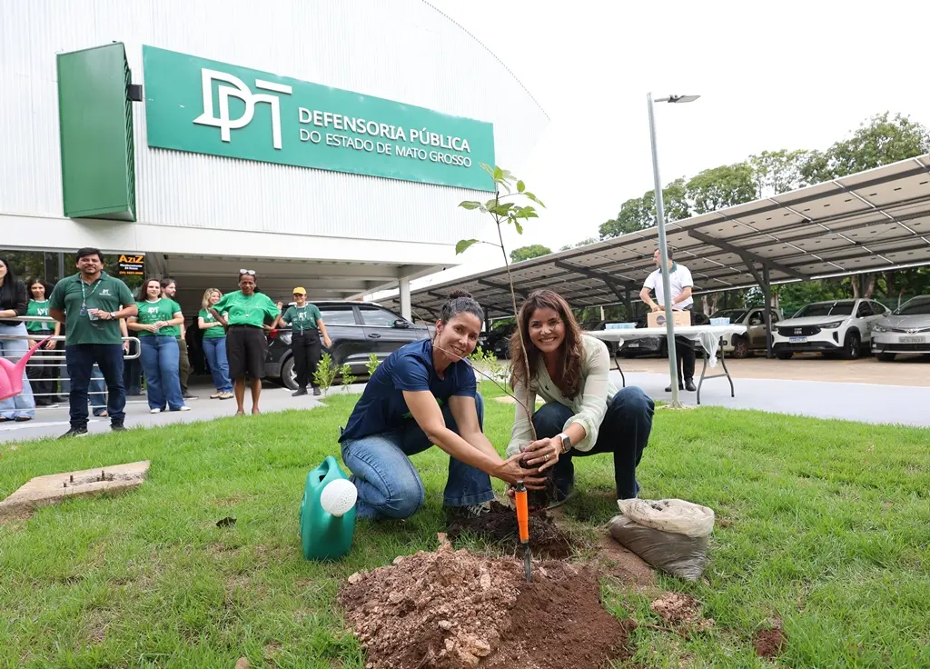 Foto horizontal colorida, em plano aberto, que mostra a engenheira florestal do programa Verde Novo, Rosiani Carnaíba, e a defensora pública-geral do Estado, Luziane Castro, sorrindo para a foto, durante plantio de uma muda de ipê, ao lado do prédio da Defensoria Pública. 