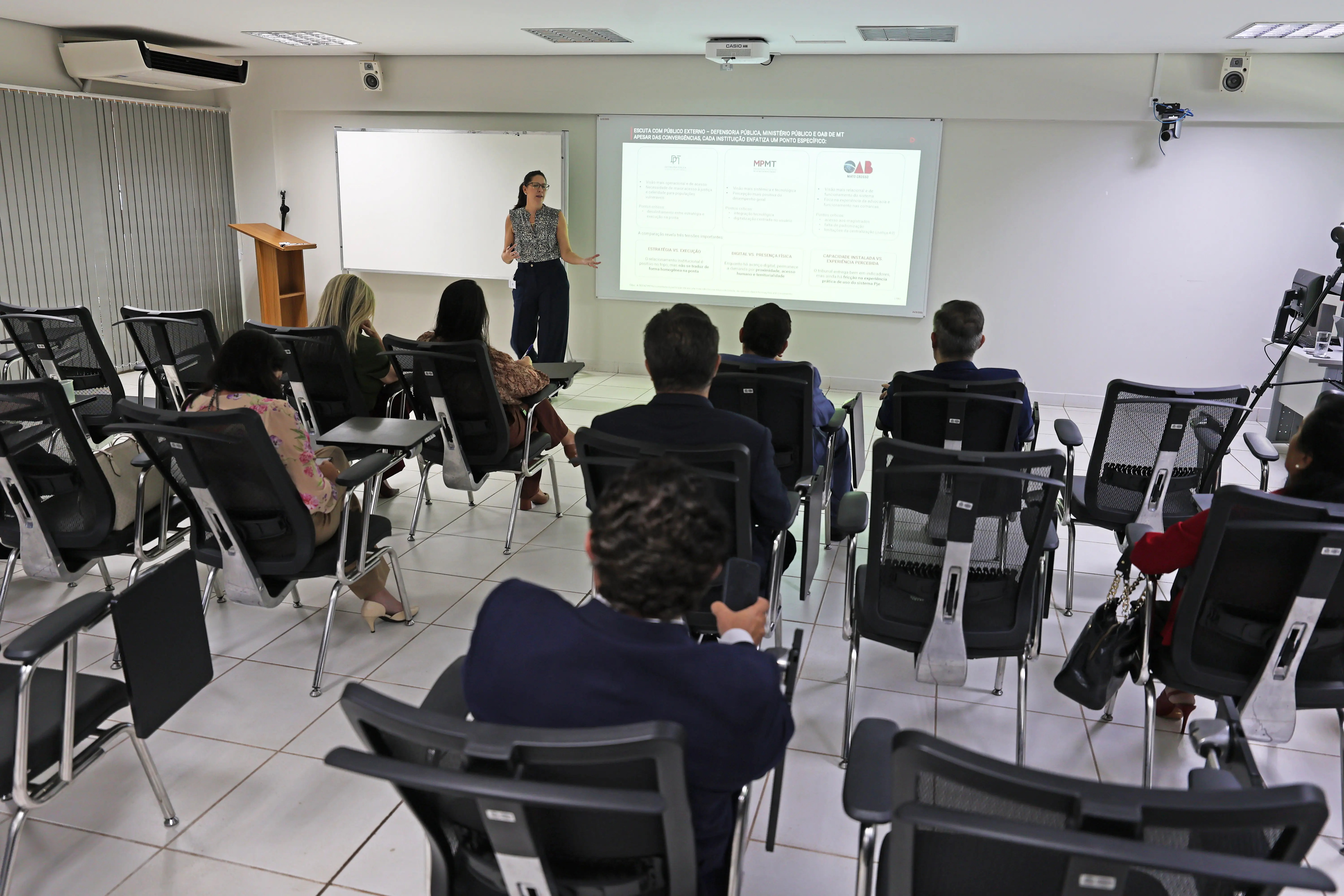 Vista panorâmica de sala de aula com pessoas sentadas de costas em cadeiras pretas. À frente, uma mulher apresenta slides em uma tela branca para o grupo.