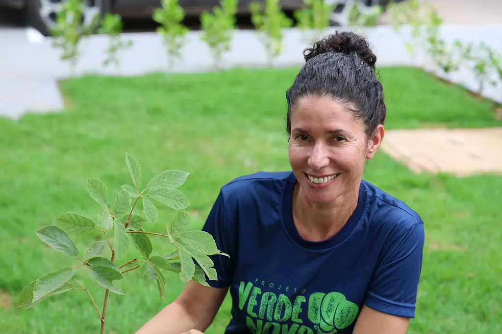 Foto horizontal colorida, em plano fechado, que mostra a engenheira florestal do programa Verde Novo, Rosiani Carnaíba, sorrindo para a foto, agachada ao lado de uma muda de árvore em um gramado. Ela é uma mulher de pele clara, magra, de cabelo preto preso em coque, usando camiseta do Verde Novo.