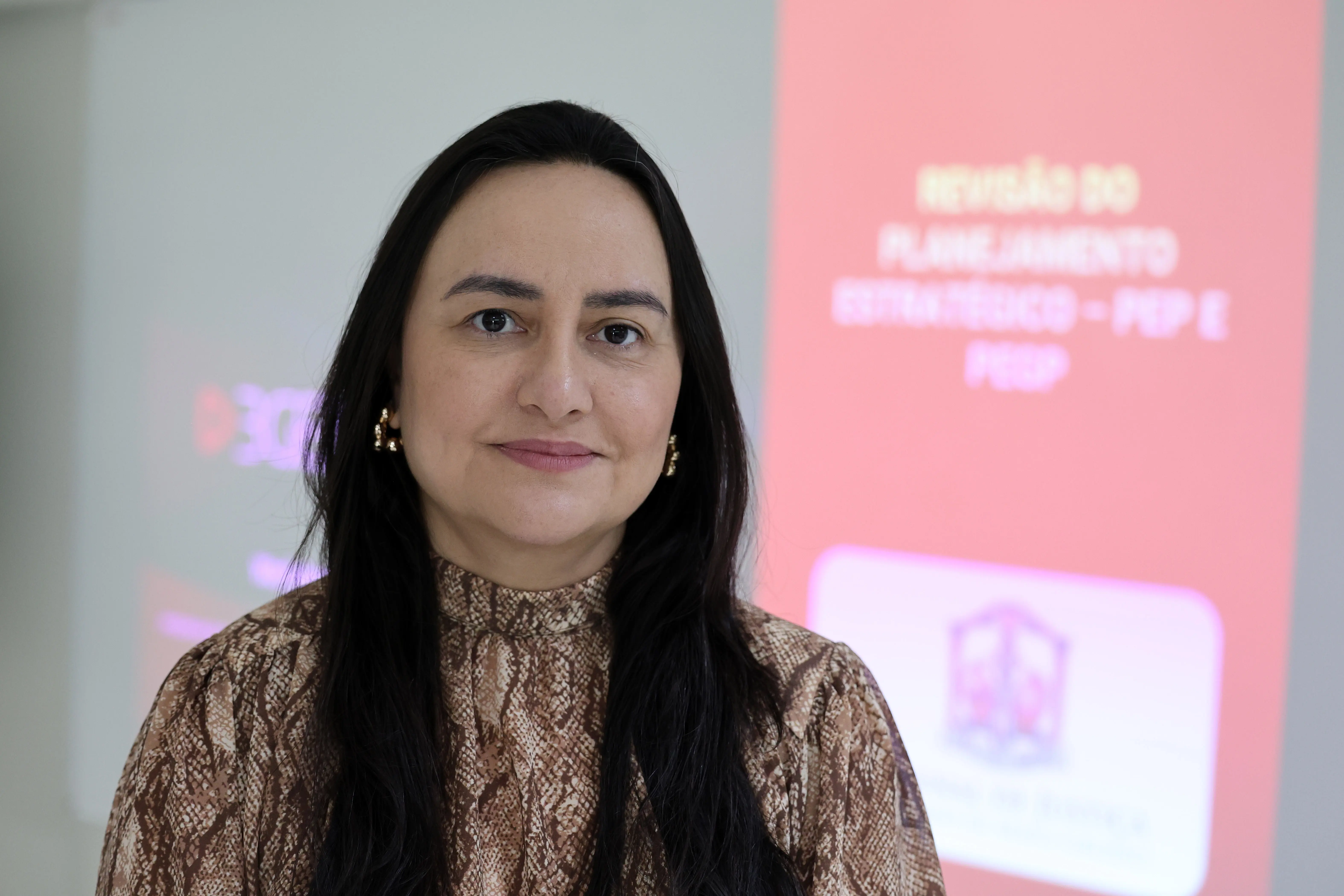 Retrato da juíza Alethea Assunção Santos. Ela tem cabelos longos escuros e blusa de estampa animal. Ao fundo, um banner rosa com o texto "Revisão do Planejamento Estratégico".