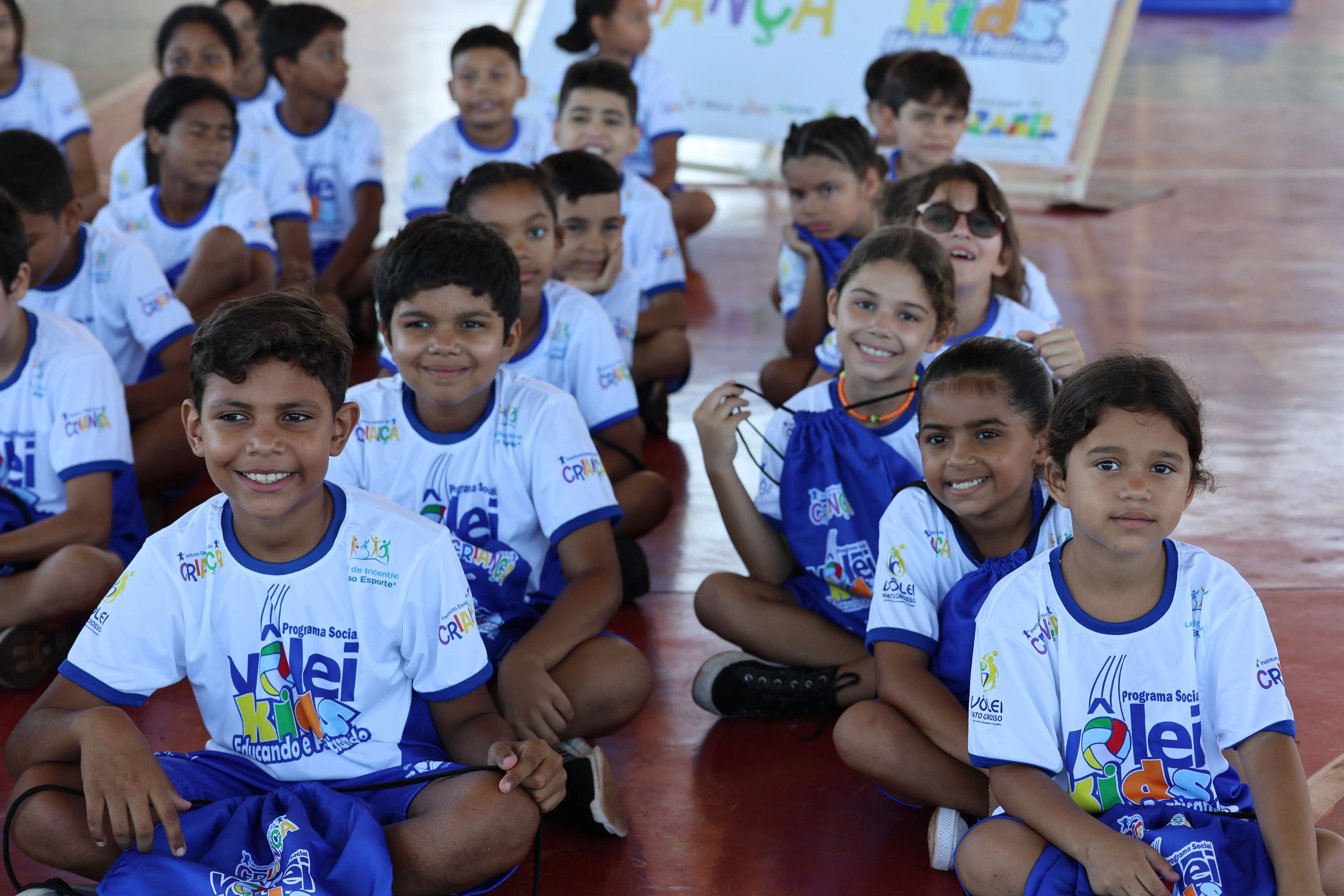 Grupo de meninos com uniforme branco e azul posa junto, sorrindo. Estão próximos uns dos outros, em ambiente escolar.