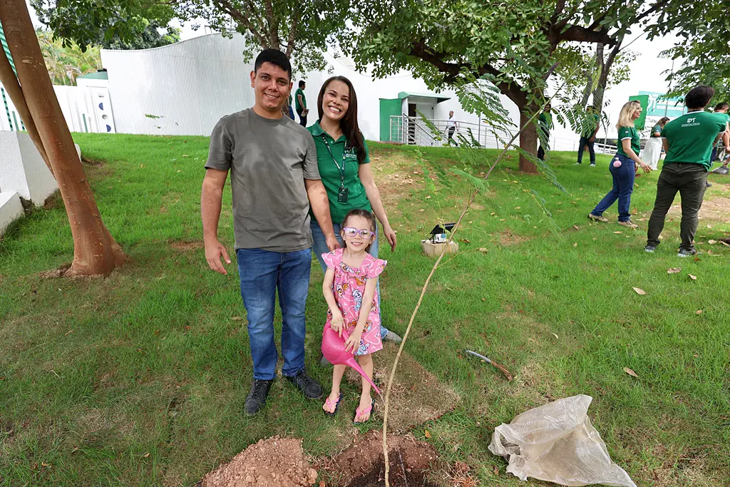 Foto horizontal colorida em plano aberto que mostra um homem, uma mulher com a camiseta da Defensoria Pública e uma menininha posando para a foto ao lado de uma muda de árvore que plantaram ao lado do prédio da Defensoria Pública. Eles sorriem para a foto.
