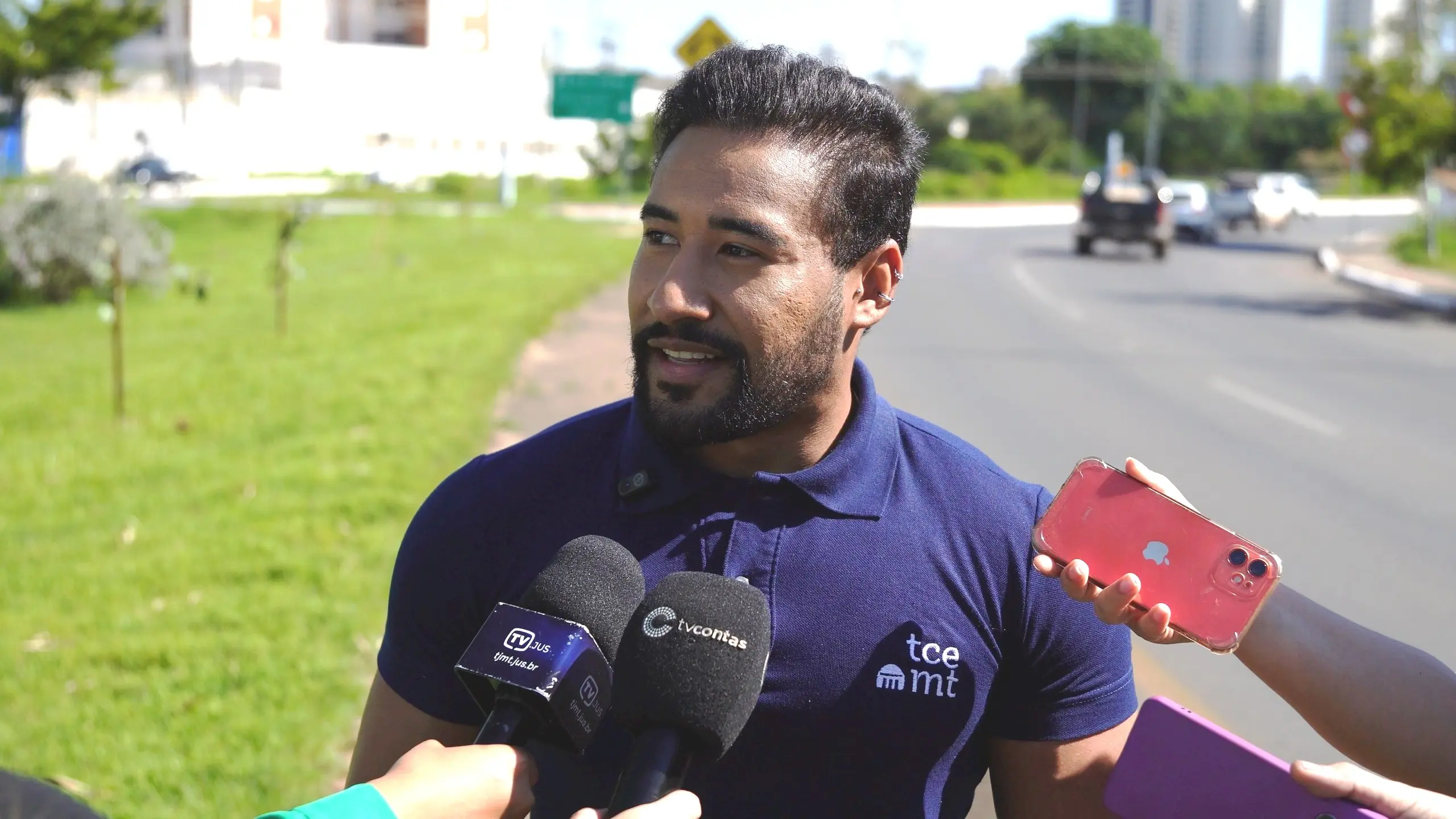 Um homem jovem, de barba e polo azul marinho com logo "tce mt", sorri enquanto fala, olhando para a esquerda. Ele é cercado por microfones e um celular vermelho que o grava. A cena se passa em uma rua com gramado e carros ao fundo.