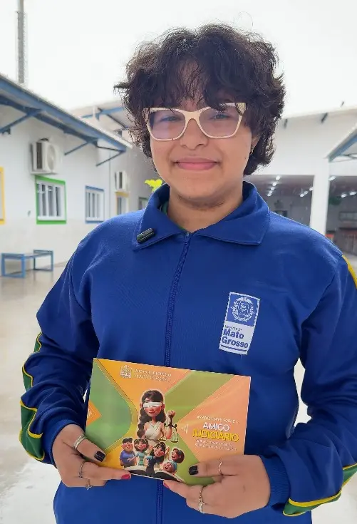 Foto vertical que mostra a aluna Manuela Sousa, de 12 anos, da cintura pra cima, no pátio da escola, sorrindo e mostrando a cartilha do Nosso Judiciário. Ela é uma menina parda, de cabelos castanhos e curtos, usando moletom azul da escola e óculos de grau. 
