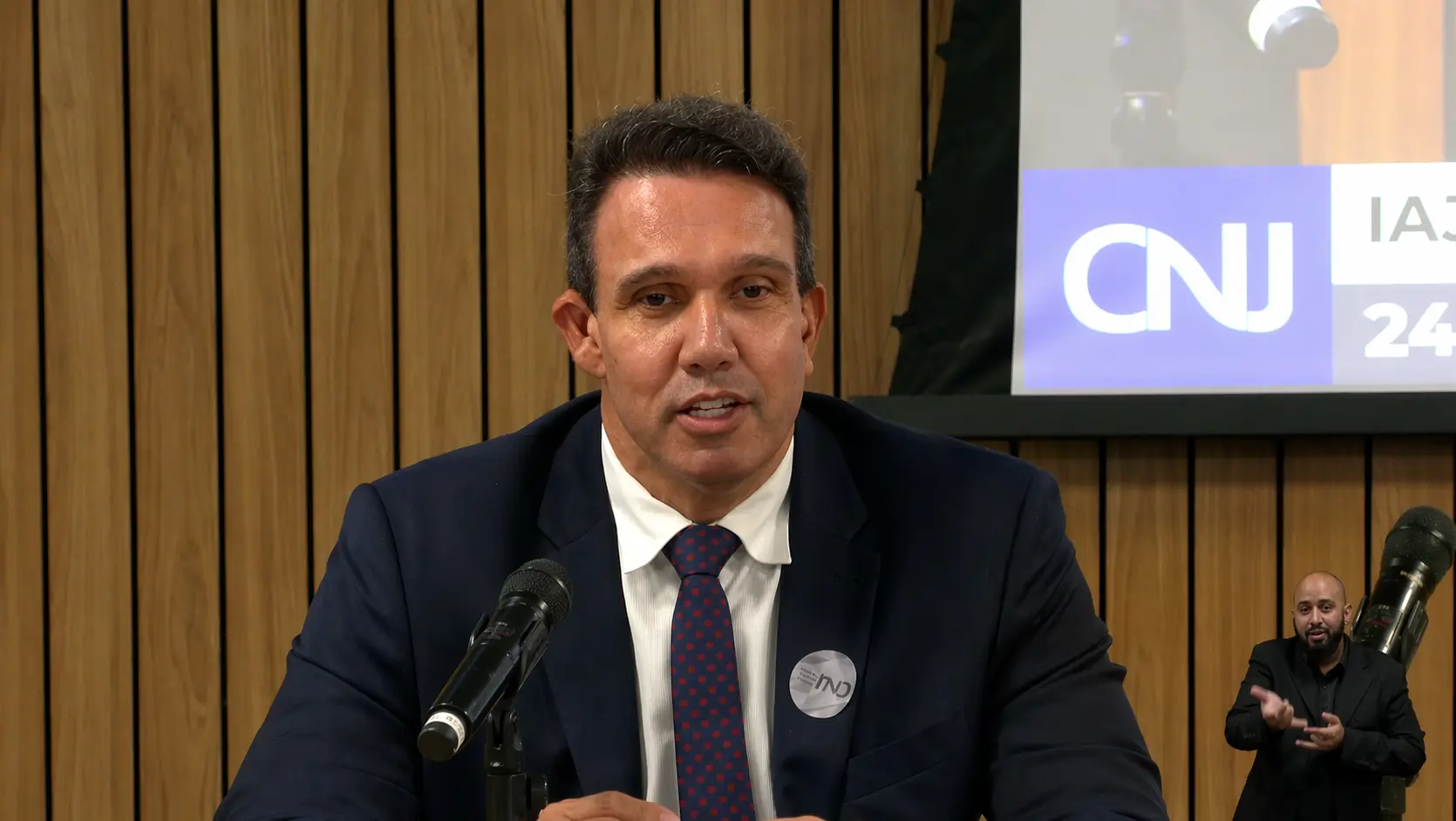 Um homem de terno fala ao microfone em um ambiente formal, com fundo de painéis de madeira e um telão com o logo do CNJ. No canto inferior direito, um intérprete de Libras faz a tradução.