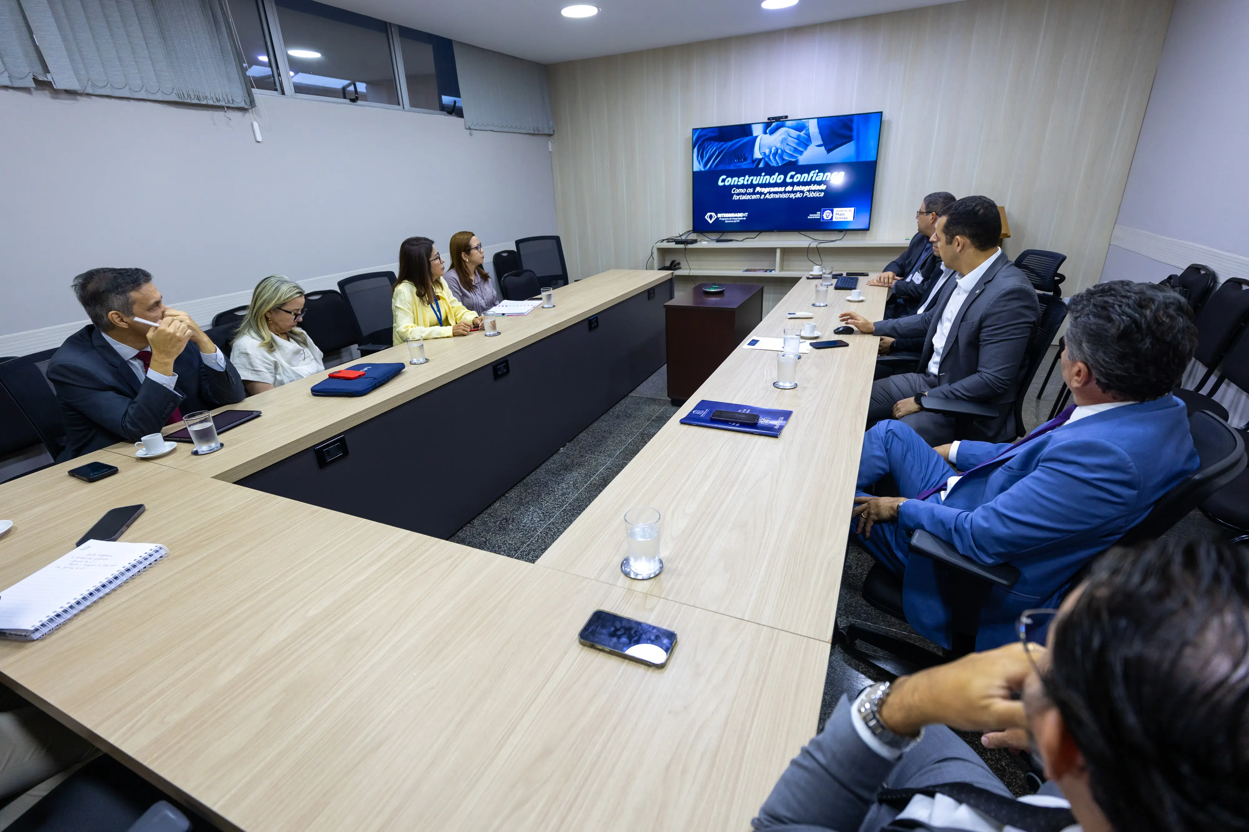 Reunião entre CGE e TJMT em sala com mesa em "U". Sete pessoas assistem a uma apresentação em TV com o título "Construindo Confiança", sobre programas de integridade na administração pública.