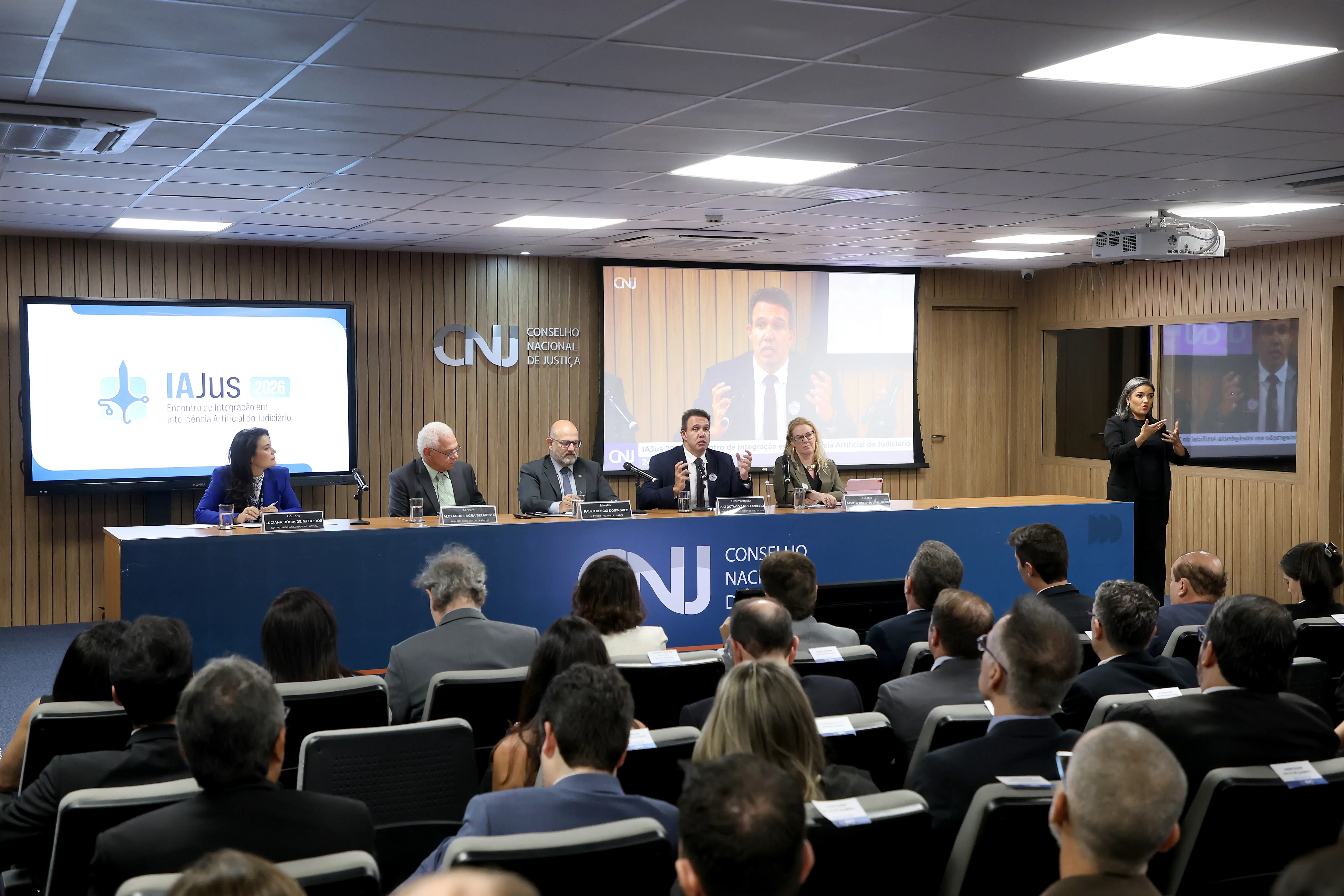 Auditório do CNJ com cinco palestrantes à mesa do IAJus 2026. Um homem fala ao centro enquanto uma intérprete de Libras sinaliza à direita. Telas exibem o logo e o palestrante para a plateia.