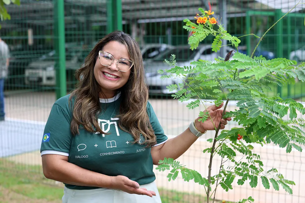 Foto horizontal colorida, em plano médio, que mostra a servidora da Defensoria Pública, Tereza Cristina Sales, sorrindo para a foto ao lado de uma muda de flamboyant que plantou ao lado do prédio da Defensoria. Ela é uma mulher negra, de cabelos longos e castanhos, usando camiseta da Defensoria.