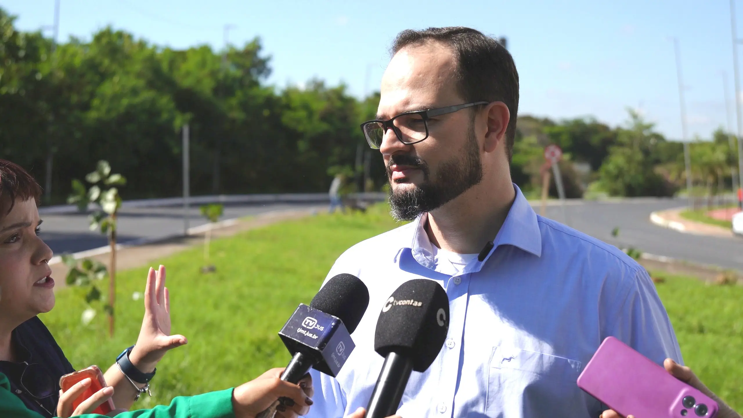 Homem jovem, de barba, óculos e camisa social azul clara, fala olhando para a esquerda. Ele é rodeado por microfones e um celular rosa. O ambiente é ao ar livre, com um gramado verde e árvores ao fundo.