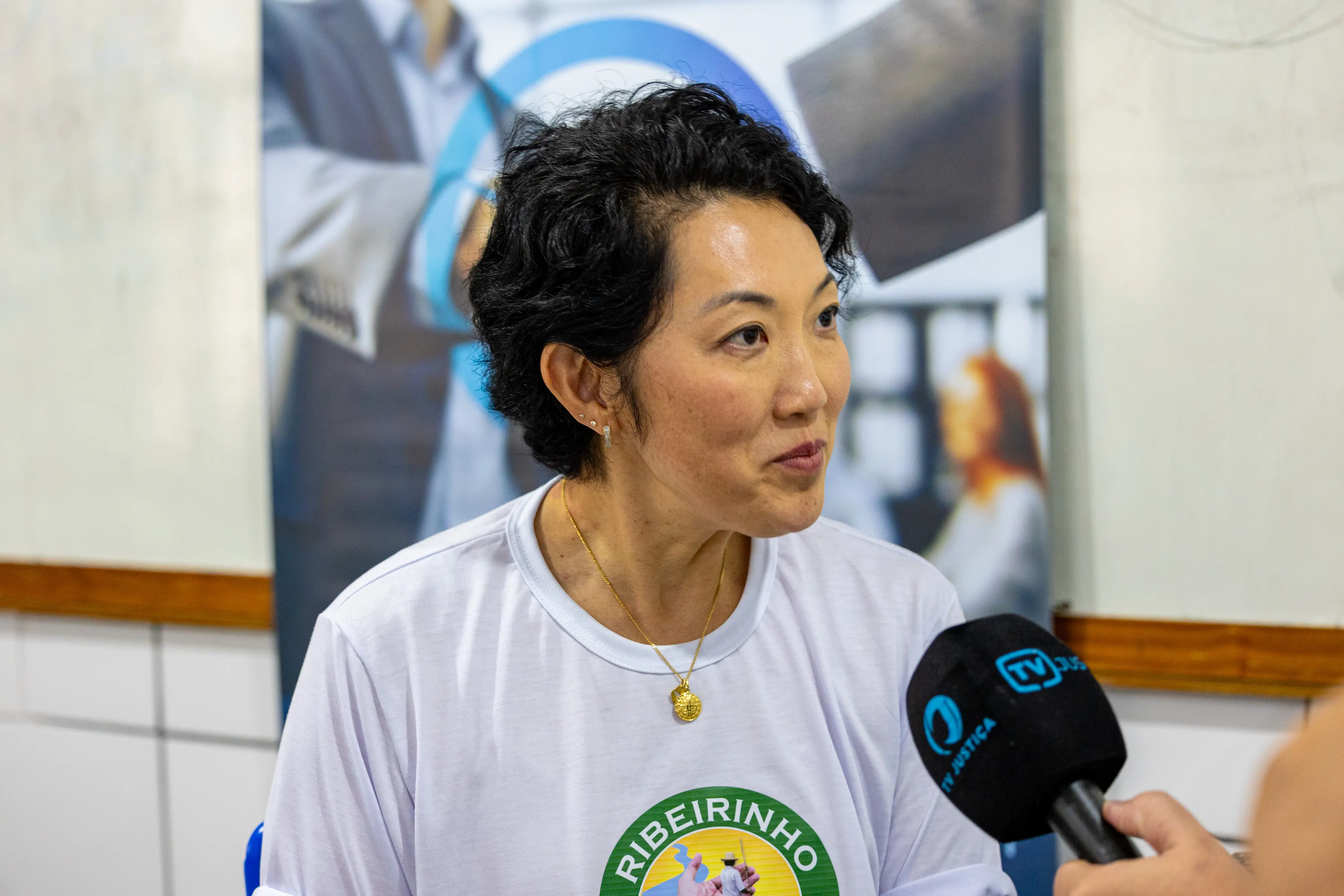 Mulher de ascendência asiática, cabelos curtos e escuros, veste camiseta branca do projeto Ribeirinho Cidadão. Ela concede entrevista ao microfone da TV Justiça em um ambiente interno.