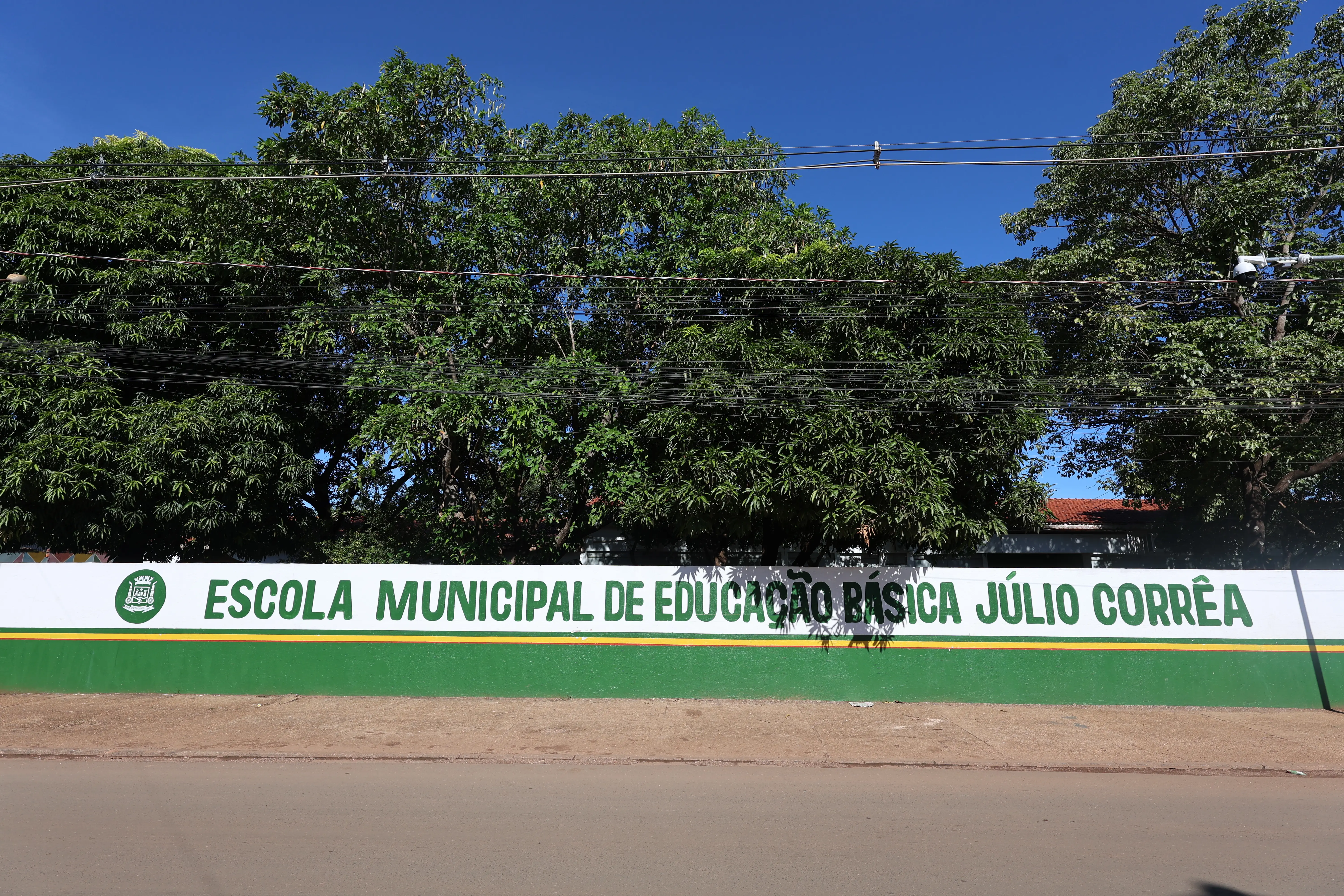 Fachada de escola municipal com placa verde e branca. Prédio baixo, cercado por árvores, em área externa durante o dia.