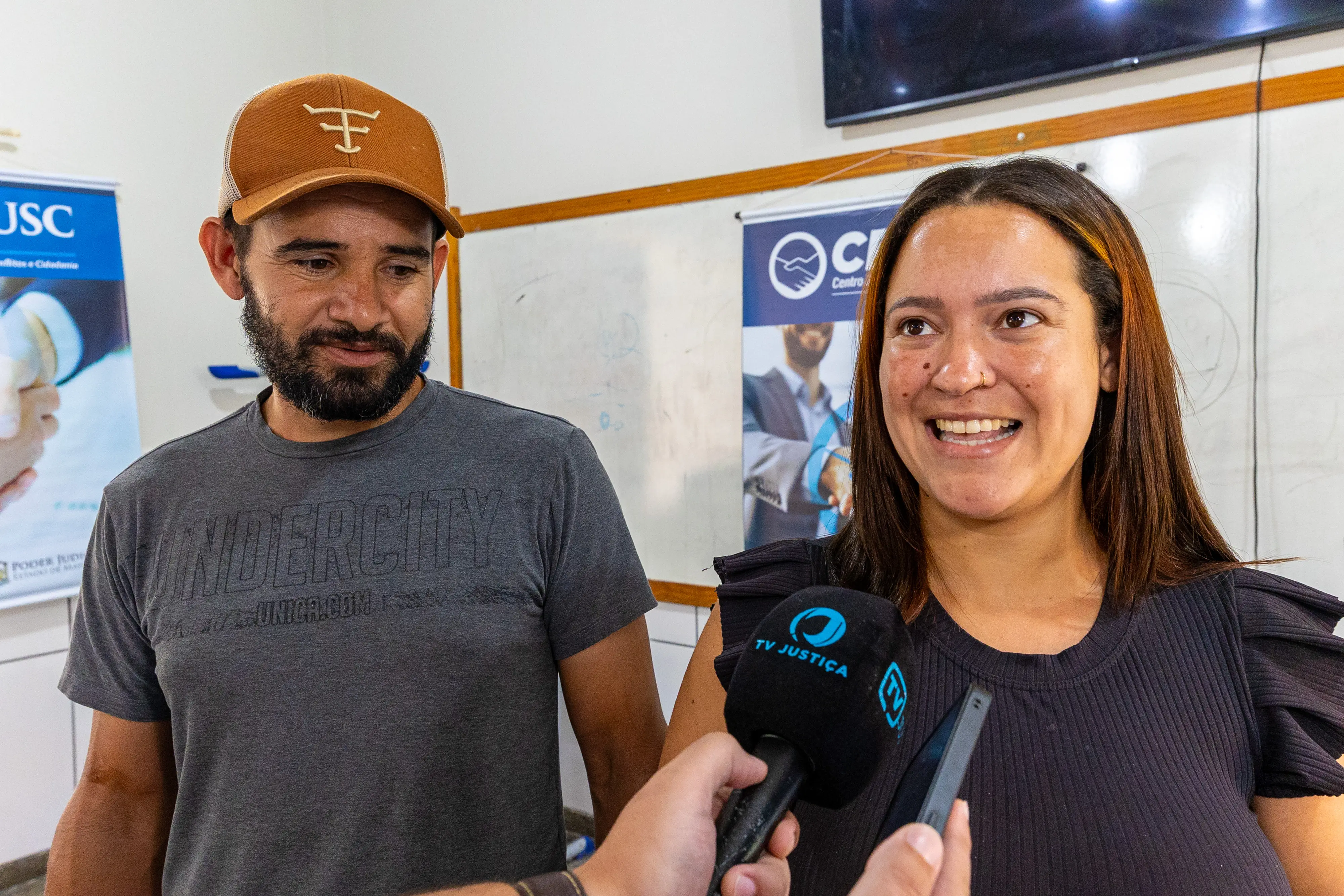 Mulher sorridente de blusa preta e homem de boné caramelo e barba concedem entrevista à TV Justiça. Ao fundo, banners do CEJUSC e um quadro branco em ambiente de atendimento.