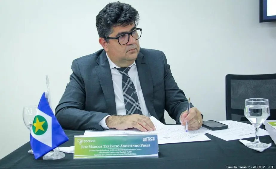 Foto horizontal do juiz Marcos Terêncio sentado e manuseando documentos sobre a mesa, durante reunião do Cocevid. Ele é um homem branco, de cabelo preto, usando camisa branca, terno cinza, gravata cinza xadrez e óculos de grau. Sobre a mesa, há uma bandeirinha de Mato Grosso.