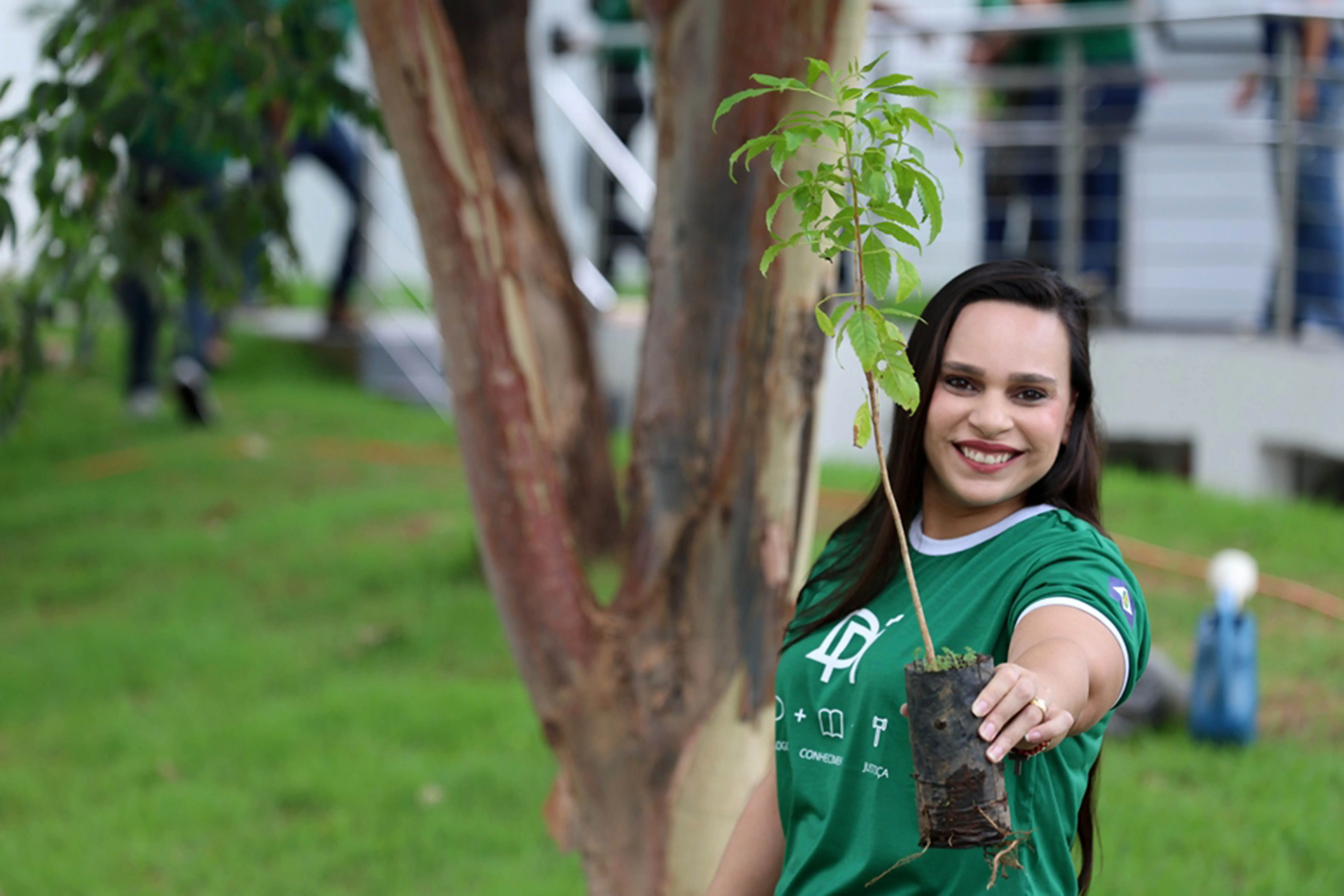 Foto horizontal colorida, em plano médio, que mostra uma servidora da Defensoria Pública Estadual, posando sorrindo para a foto e segurando uma muda de árvore. Ela é uma mulher de pele clara, cabelos longos e castanhos, usando camiseta verde da Defensoria. Atrás dela há um gramado e árvores.