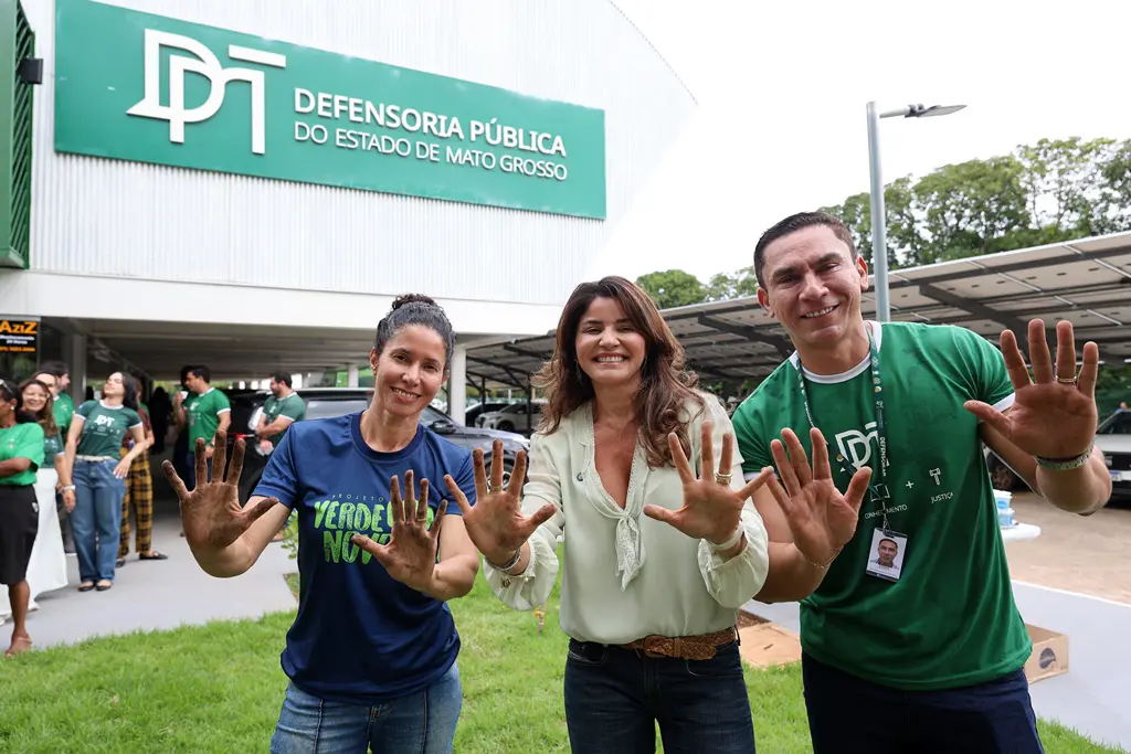 Foto horizontal colorida, em plano médio , que mostra a engenheira florestal do programa Verde Novo, a defensora pública-geral do Estado e um servidor da Defensoria Pública posando para a foto, em frente à fachada da sede da instituição. Eles sorriem e mostram as mãos sujas de terra, após plantio.