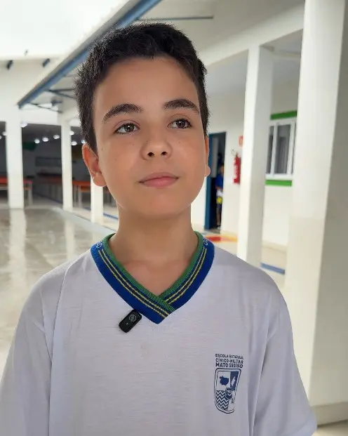 Foto vertical que mostra o aluno Kayque Wilhians, de 12 anos, da cintura pra cima, no pátio da escola. Ele é um menino branco, de olhos e cabelos castanho escuro, usando uniforme branco com detalhes azul e verde da escola. 