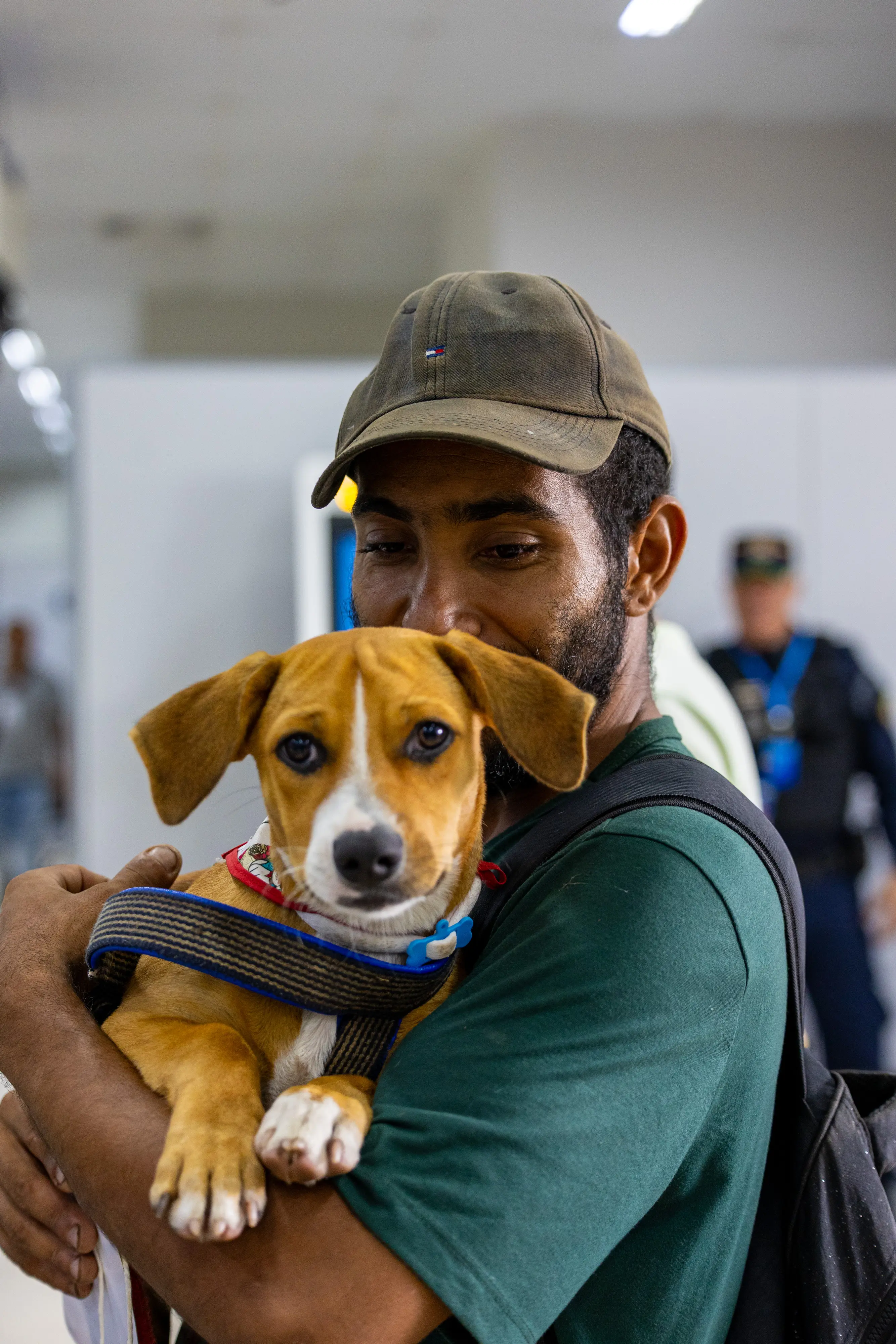 Close de um homem jovem com barba e boné marrom, segurando carinhosamente no colo um cachorro de pequeno porte, pelagem caramelo e branca, que olha fixamente para a câmera.