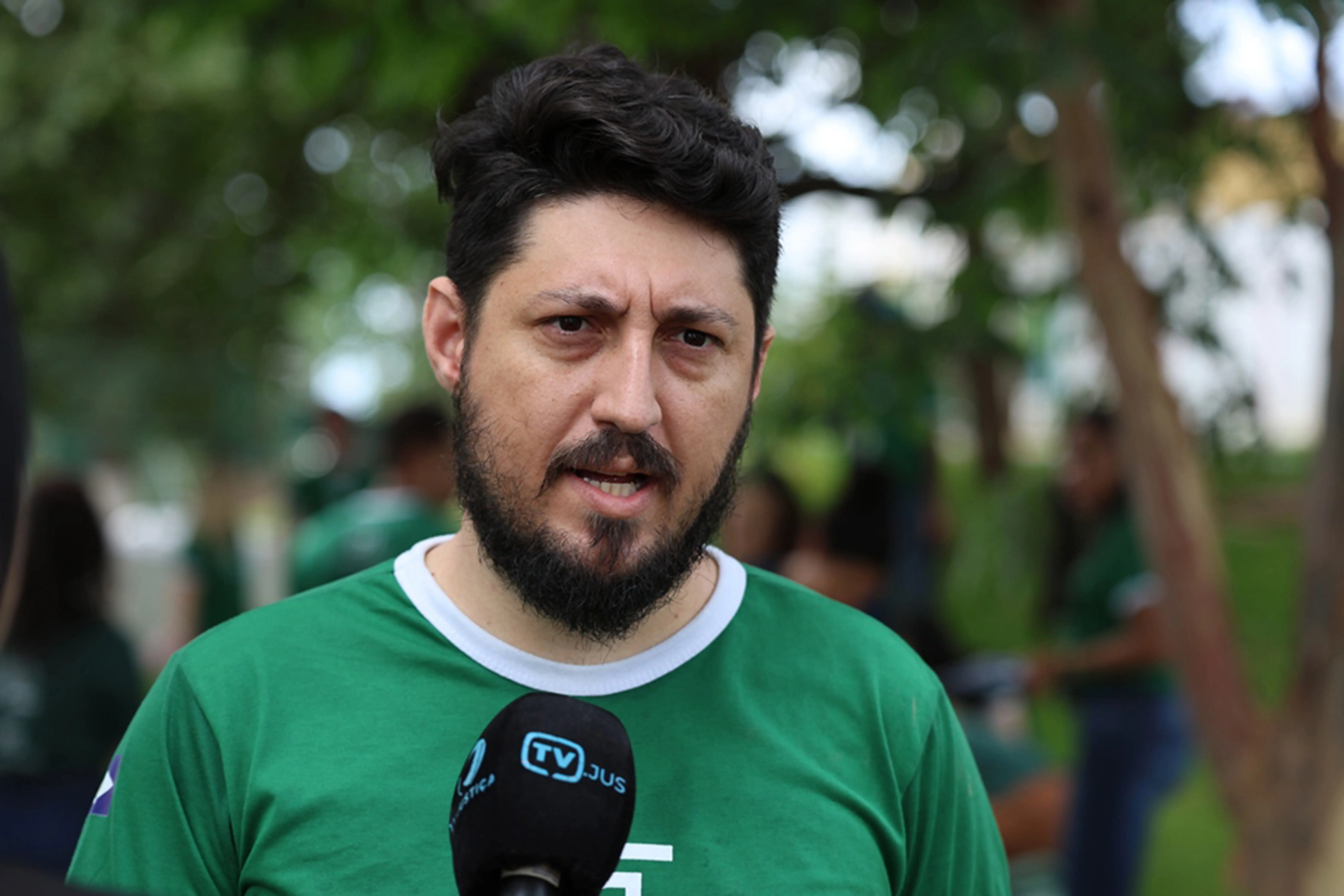 Foto horizontal colorida, em plano médio, que mostra Augusto de Souza Melo, engenheiro da Defensoria Pública, durante entrevista à TV.Jus. Ele é um homem branco, alto, de olhos, cabelos e barba escuros, usando camiseta verde da Defensoria. Atrás dele, desfocado, está um bosque e pessoas.