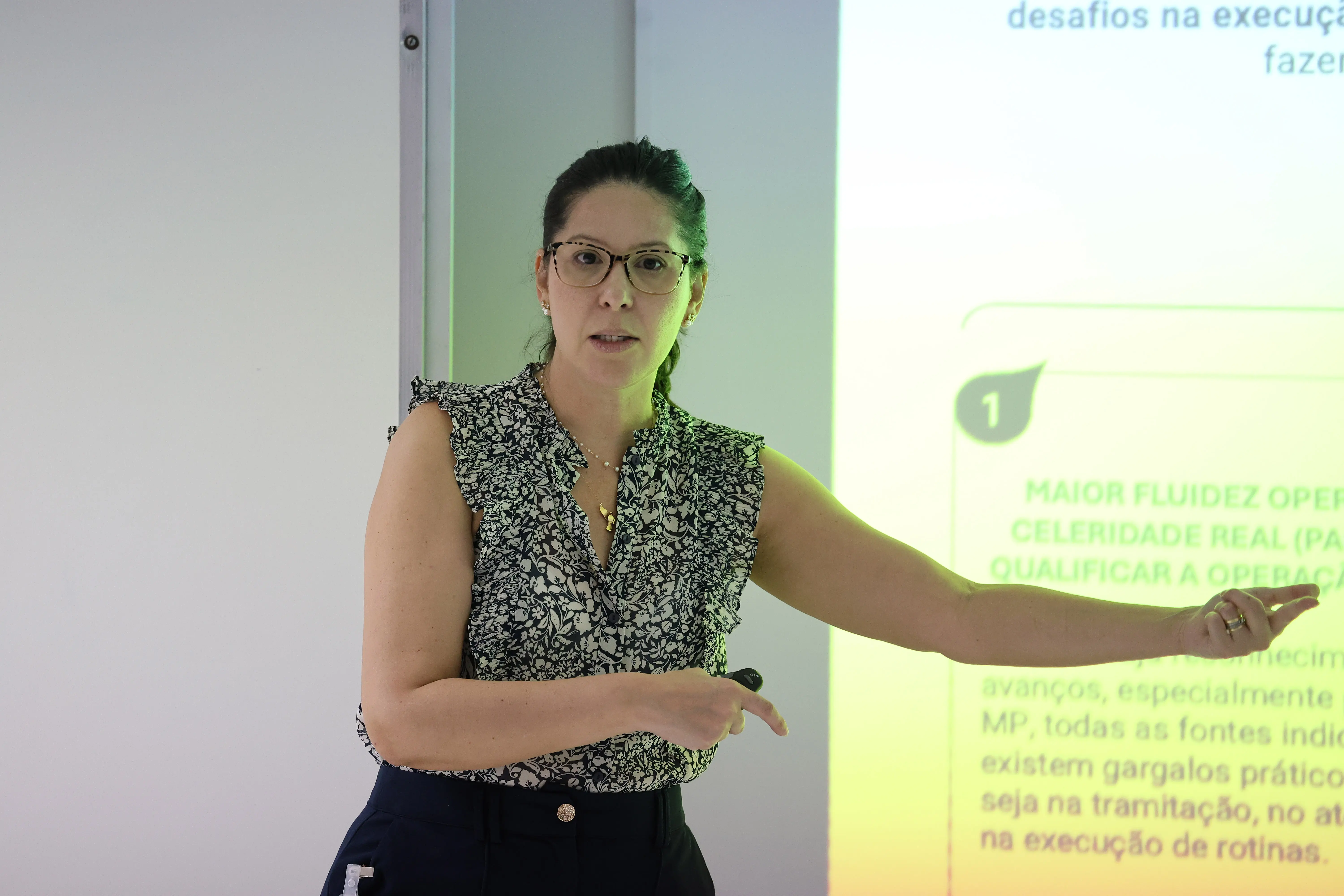 A palestrante Aline Veneri usa blusa estampada, óculos e cabelo preso. Ela gesticula à frente de uma projeção clara e aponta para o slide enquanto fala para a plateia.