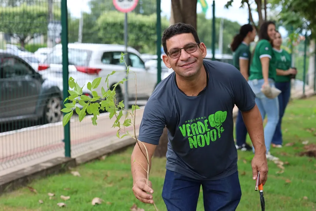 Foto horizontal colorida, em plano médio, que mostra servidor do programa Verde Novo posando para a foto ao lado de uma muda de árvores plantada em um gramado. Ele é um homem pardo, de cabelos castanhos, usando calça jeans, camiseta cinza com a logomarca do Verde Novo e óculos de grau.