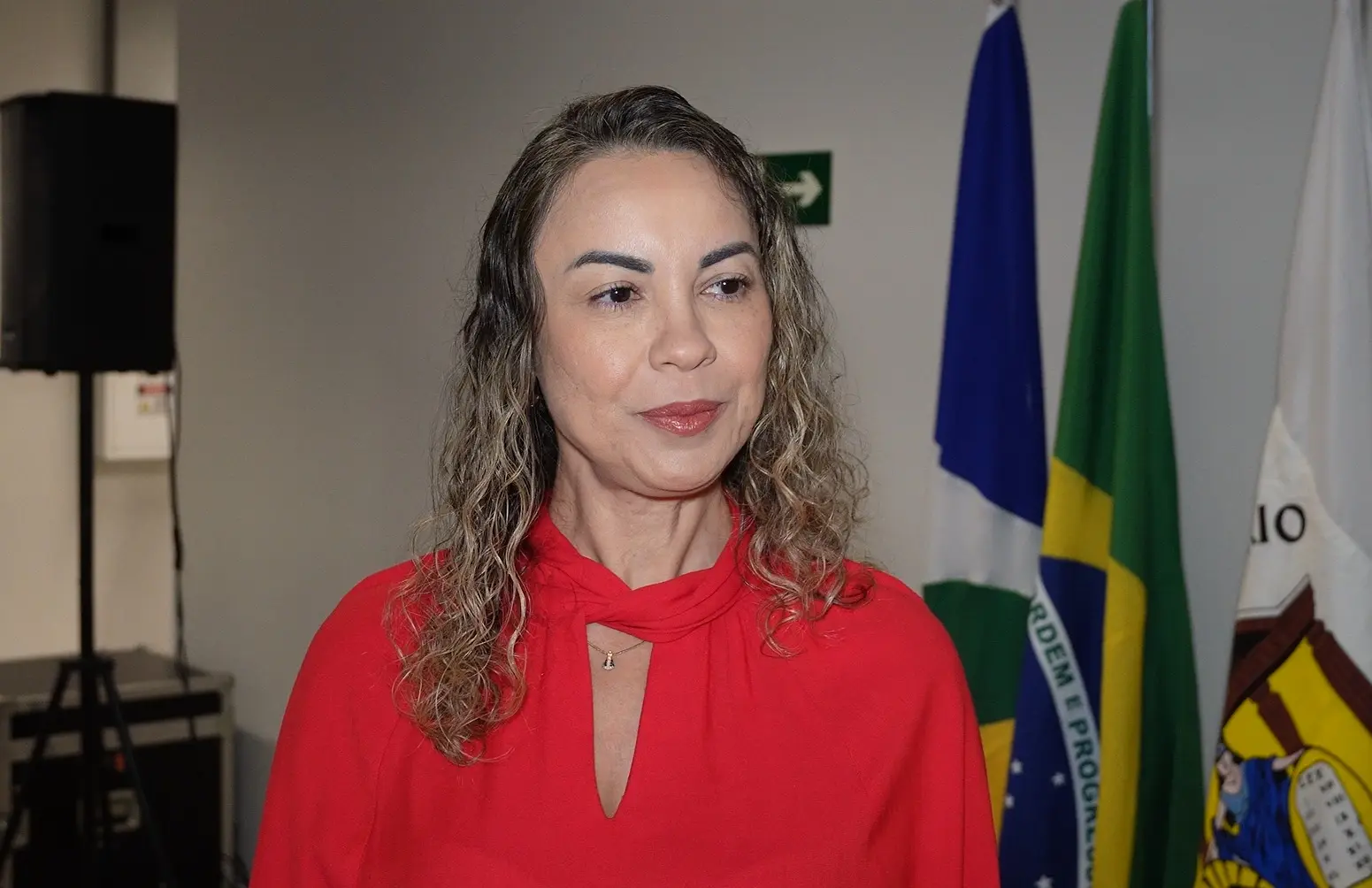 Foto horizontal que mostra a juíza Cristiane Padim, do busto pra cima, no saguão do Complexo dos Juizados. Ela é uma mulher branca, de olhos castanhos, cabelos longos, loiros e cacheados, usando blusa vermelha.
