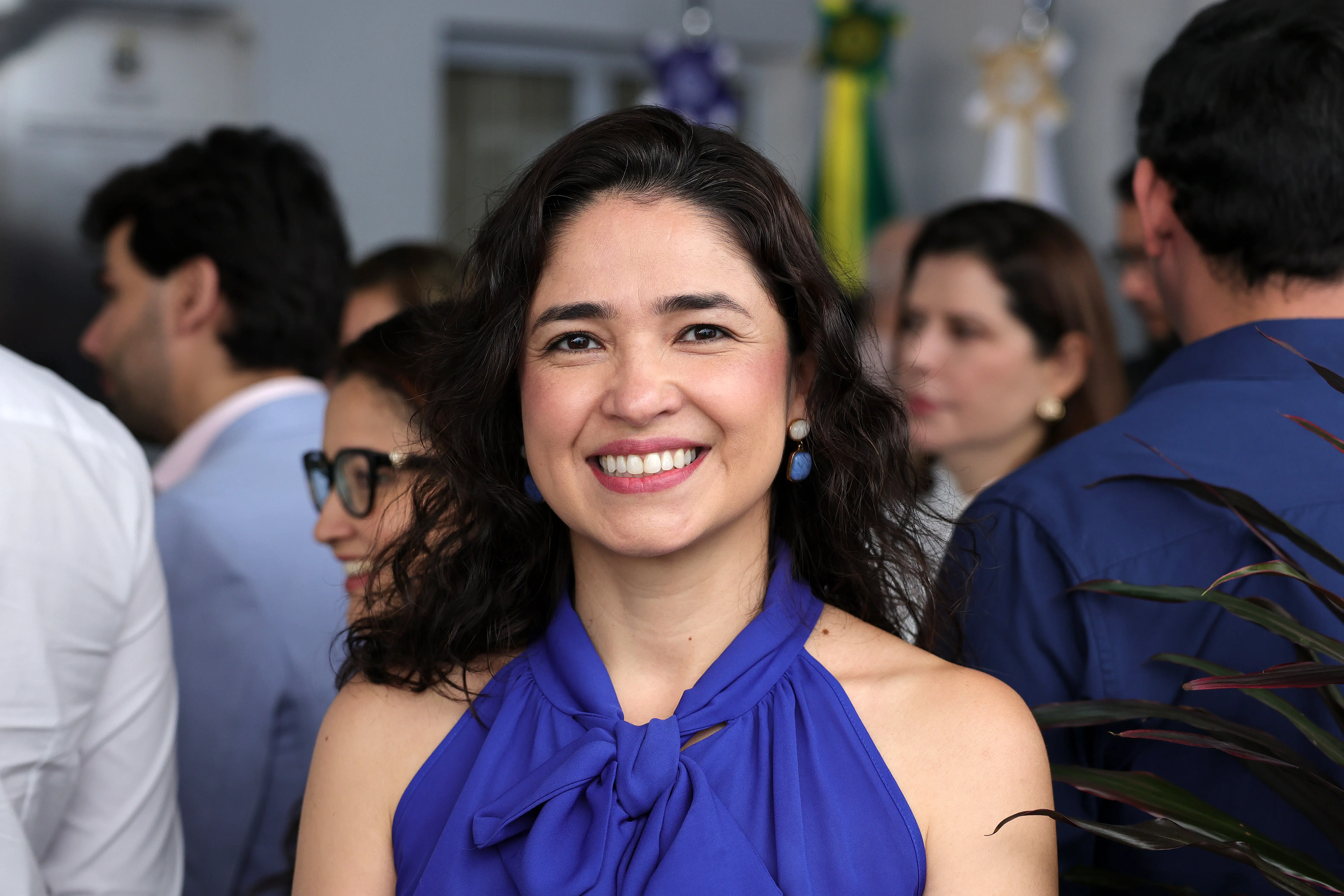 Retrato em plano médio de uma mulher sorrindo. Ela tem cabelos escuros ondulados, usa brincos azuis e blusa azul-bic com nó no decote. Há outras pessoas desfocadas ao fundo.