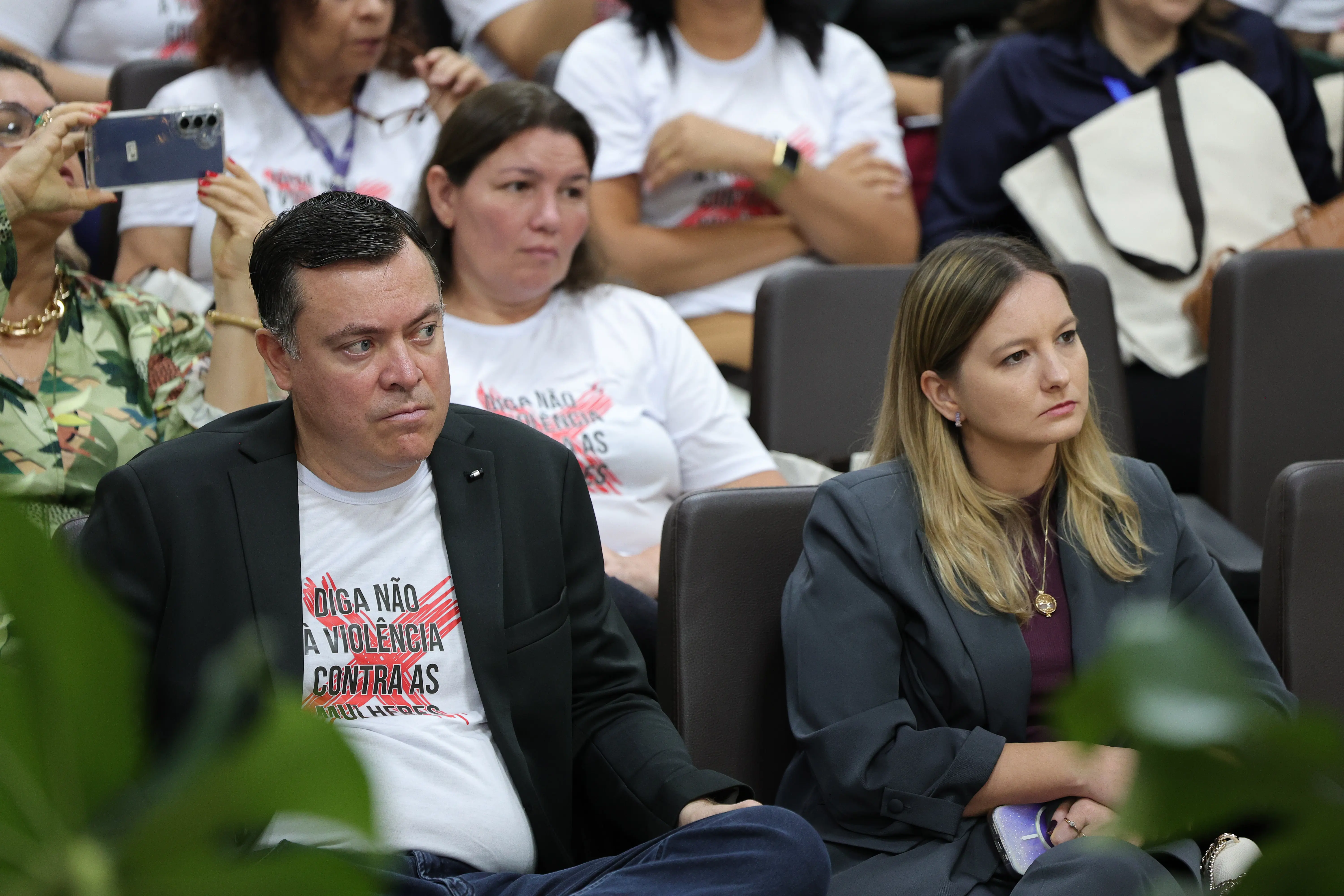 Um homem de paletó preto sobre camiseta branca da campanha e uma mulher de blazer cinza estão sentados lado a lado nas primeiras fileiras do auditório, ambos olhando sérios para o palco.