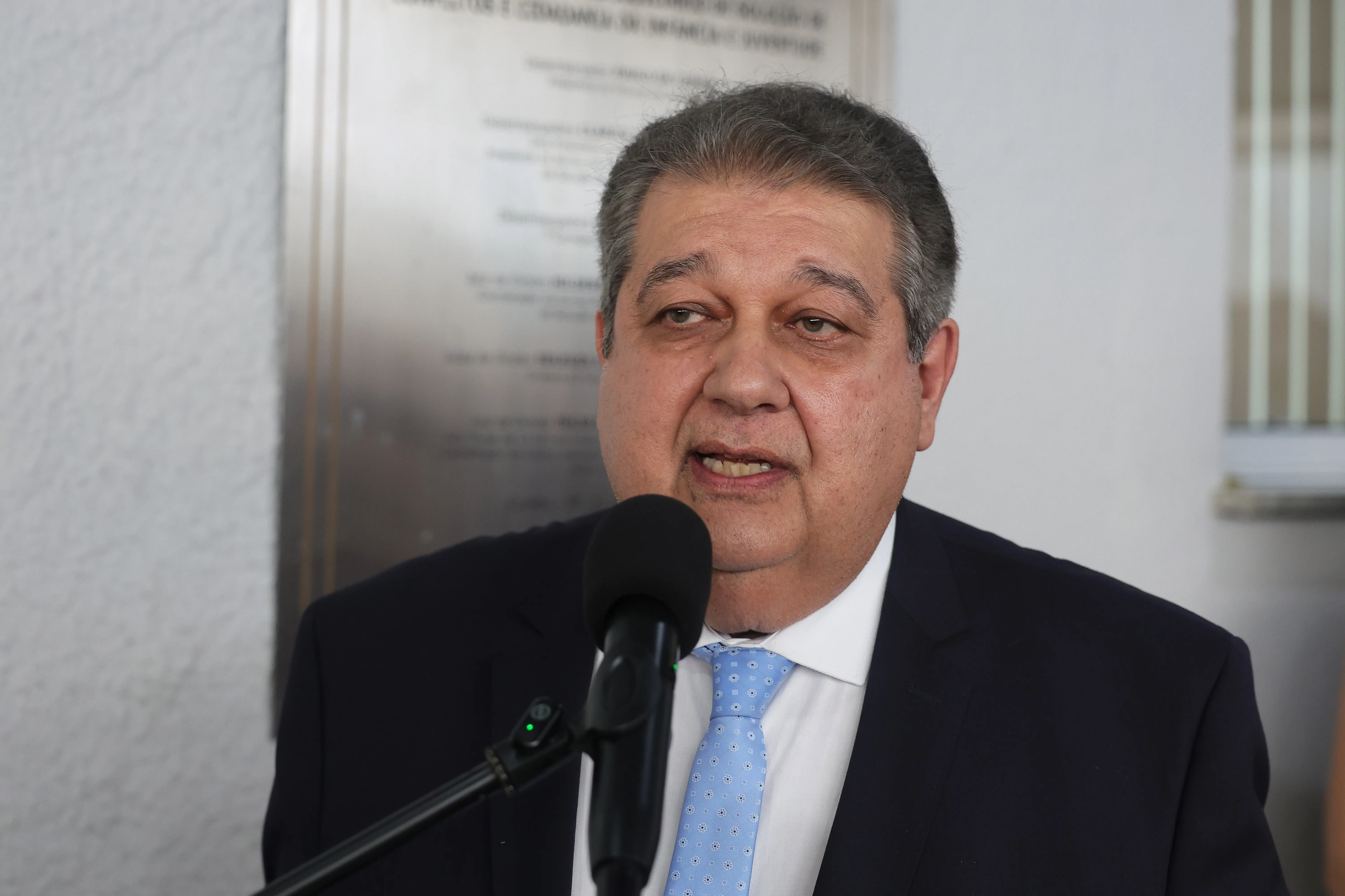 Homem de pele clara, cabelos grisalhos e terno escuro com gravata azul, falando em um microfone preto em primeiro plano. Ao fundo, há uma placa metálica fixada na parede clara.