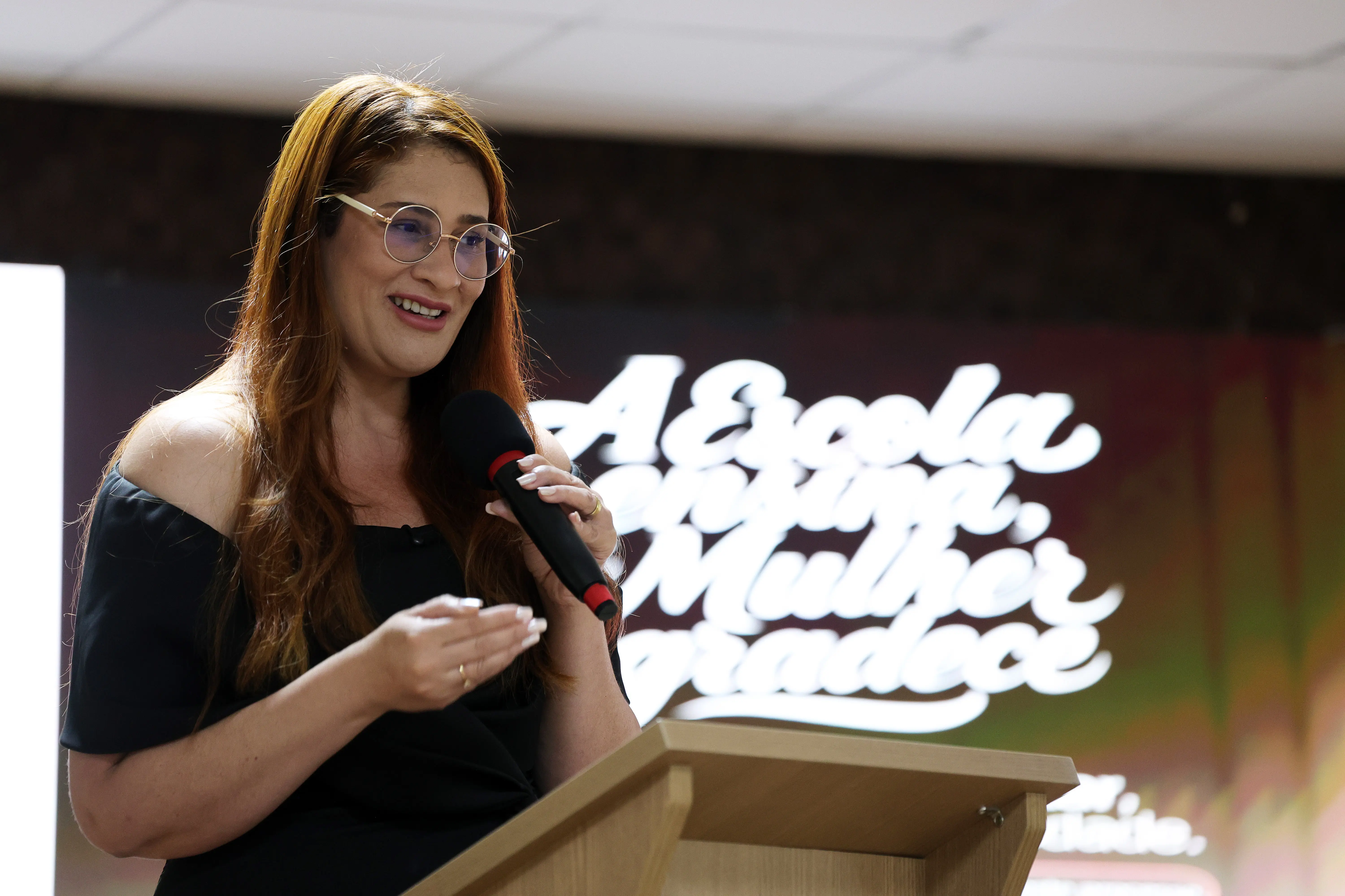 Mulher de cabelos longos acobreados e óculos de grau fala ao microfone atrás de um púlpito de madeira. Usa blusa preta de ombros de fora. Ao fundo, painel com a escrita "A Escola ensina, a Mulher agradece".