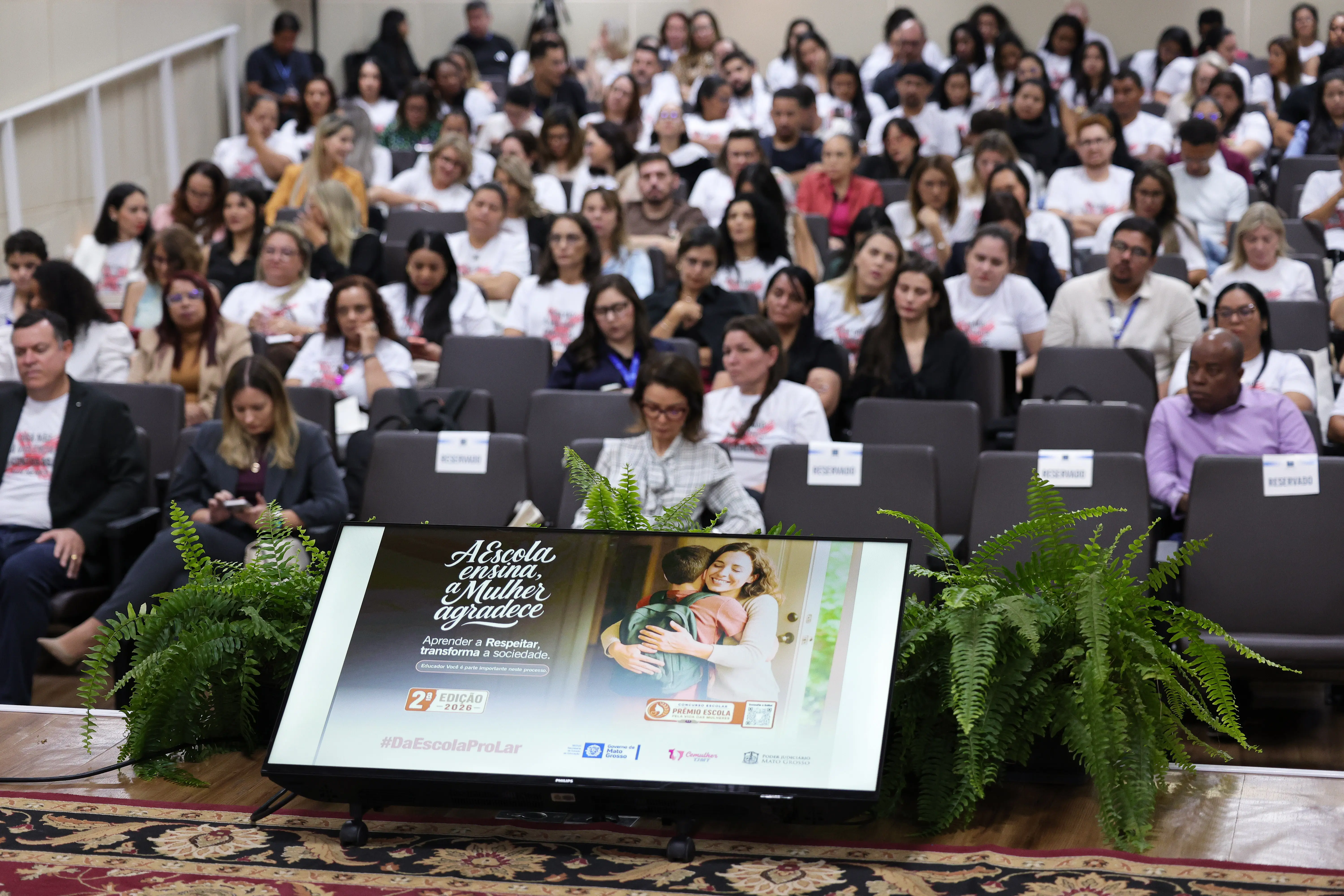 Vista do palco para o auditório com plateia lotada. Em primeiro plano, uma tela no chão exibe a imagem da campanha "A Escola ensina, a Mulher agradece" entre dois vasos de samambaias.