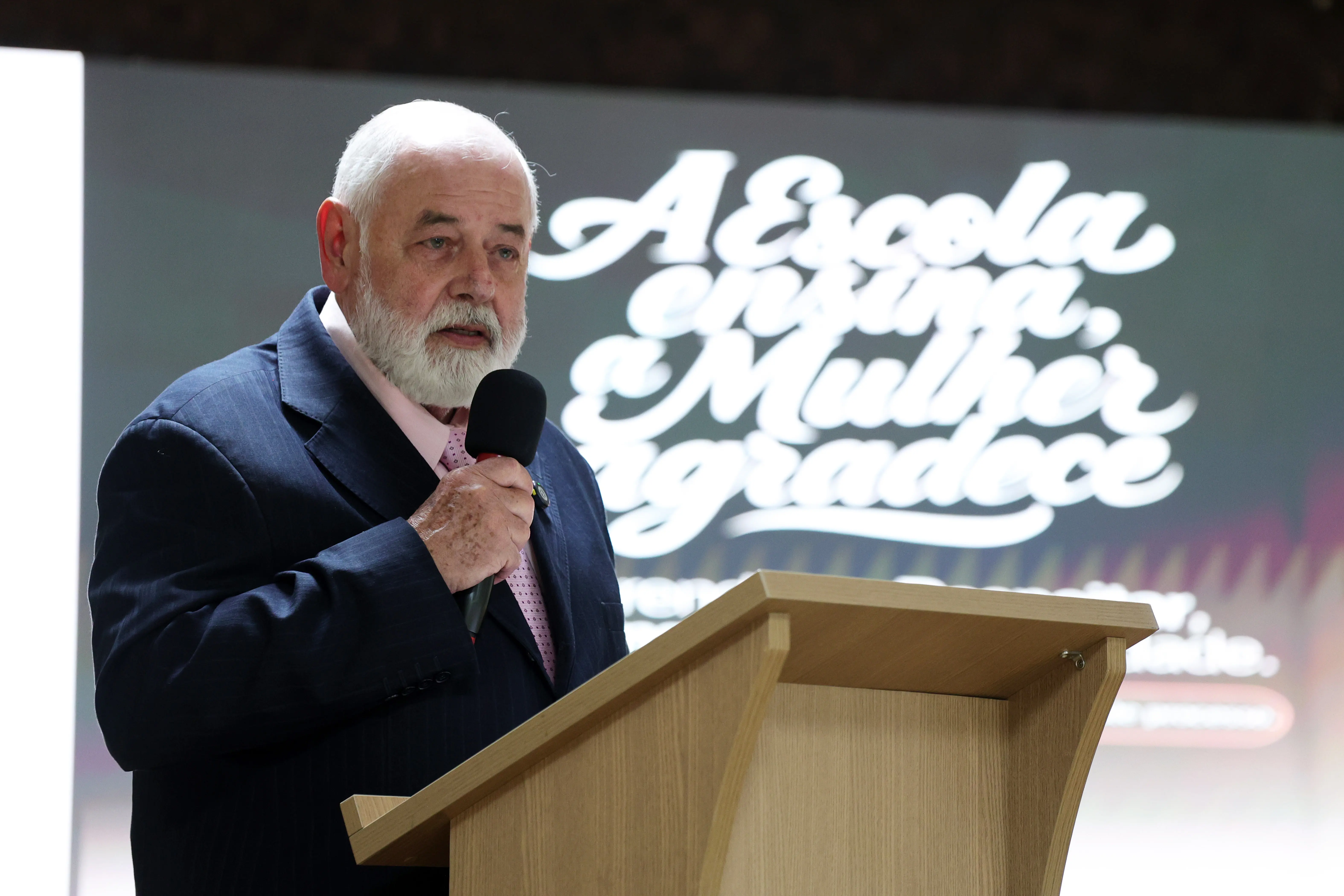 Homem idoso de cabelo e barba brancos fala ao microfone atrás de um púlpito de madeira. Usa terno azul-marinho e gravata clara. Ao fundo, painel com a frase "A Escola ensina, a Mulher agradece".