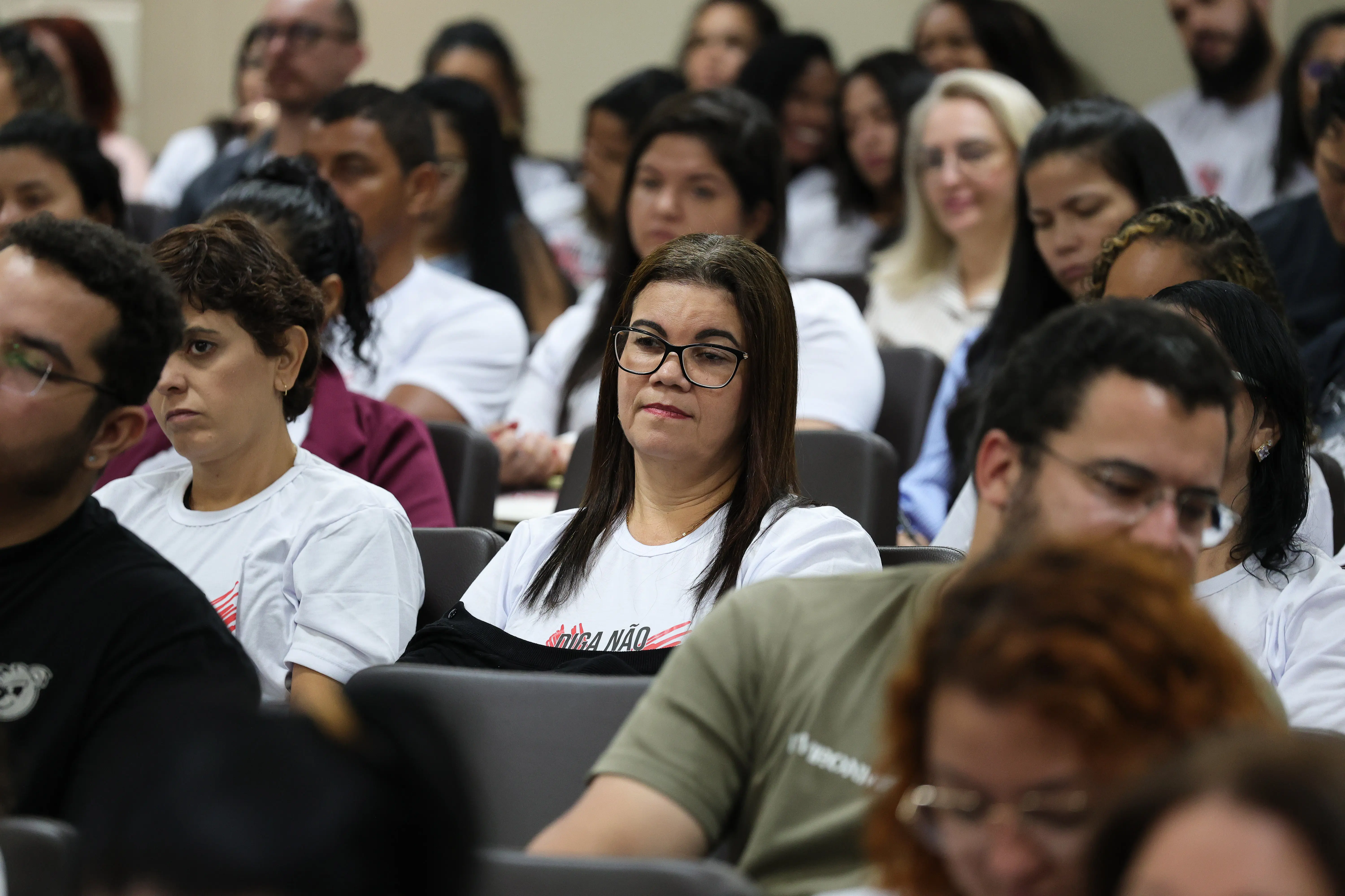 Mulher de cabelos castanhos e óculos de grau, vestindo camiseta branca com a estampa da campanha, sentada no meio da plateia e olhando com atenção para a frente.
