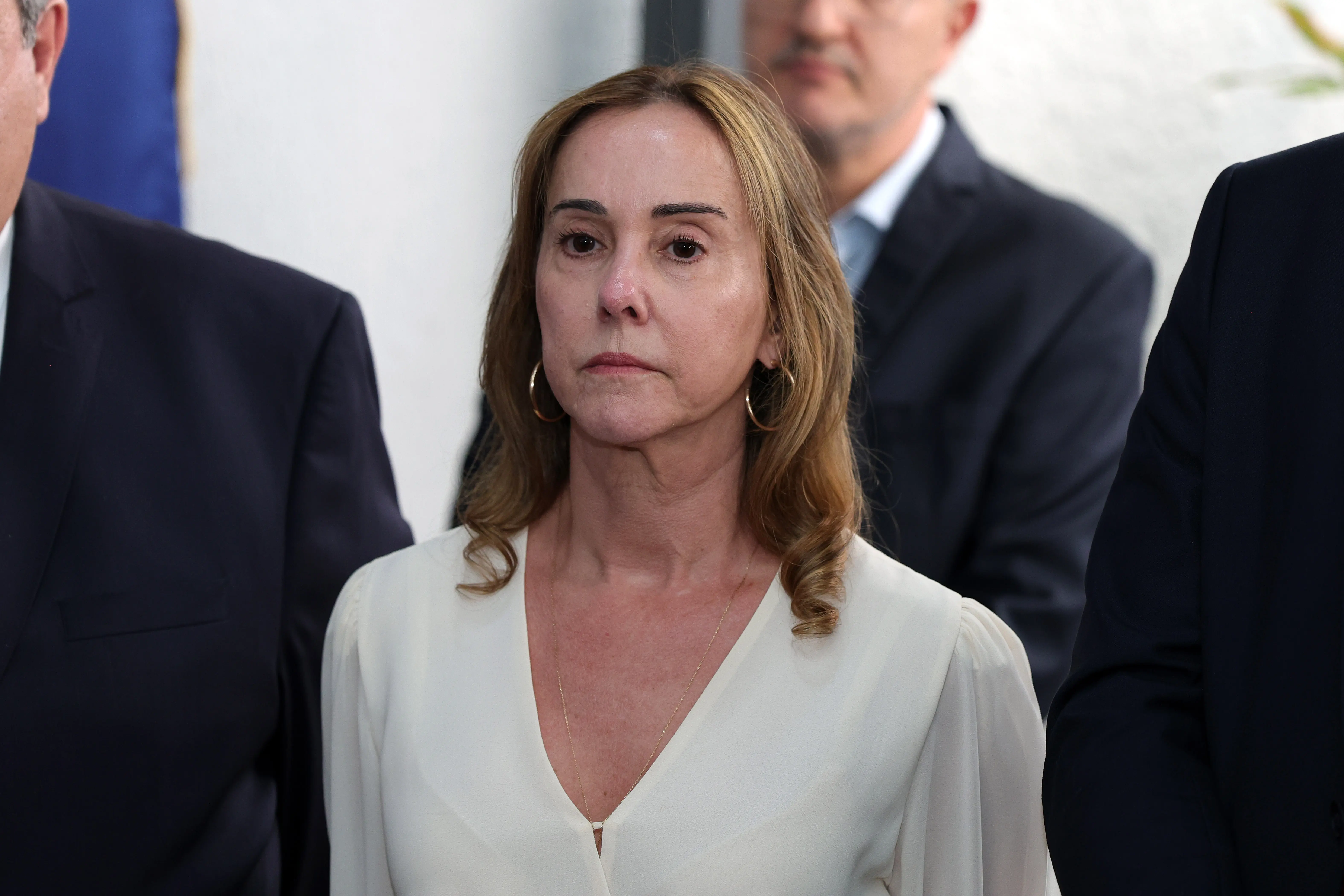 Fotografia aproximada de uma mulher de pele clara e cabelos loiros ondulados, vestindo blusa branca com decote em V e brincos de argola dourados. Homens de terno escuro estão ao seu redor.