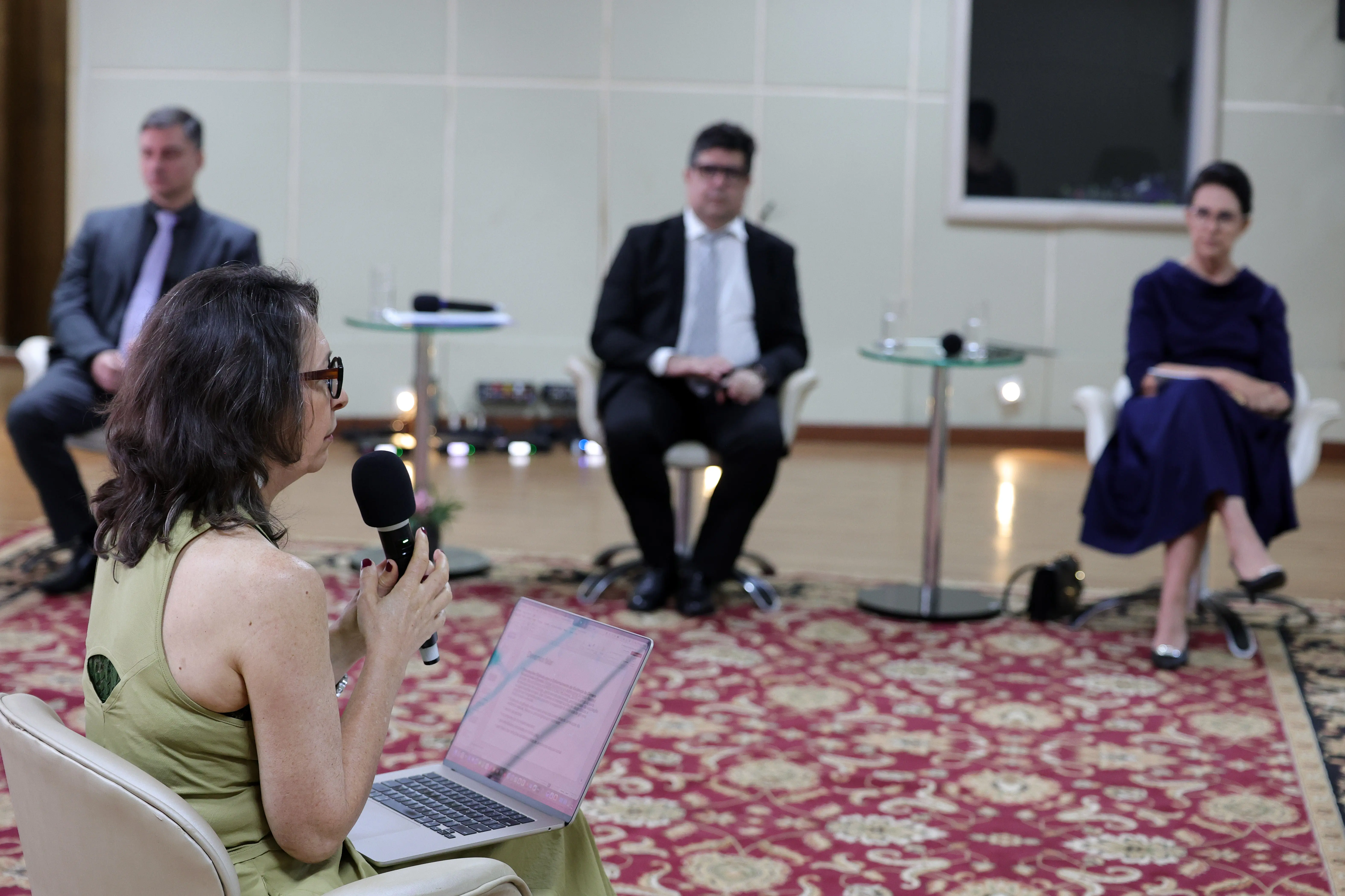 Vista em primeiro plano lateral de uma mulher no palco segurando um microfone e com notebook aberto. Ao fundo, desfocados, três participantes compostos por dois homens de terno e uma mulher de vestido azul-marinho assistem sentados em cadeiras brancas sobre um tapete estampado.