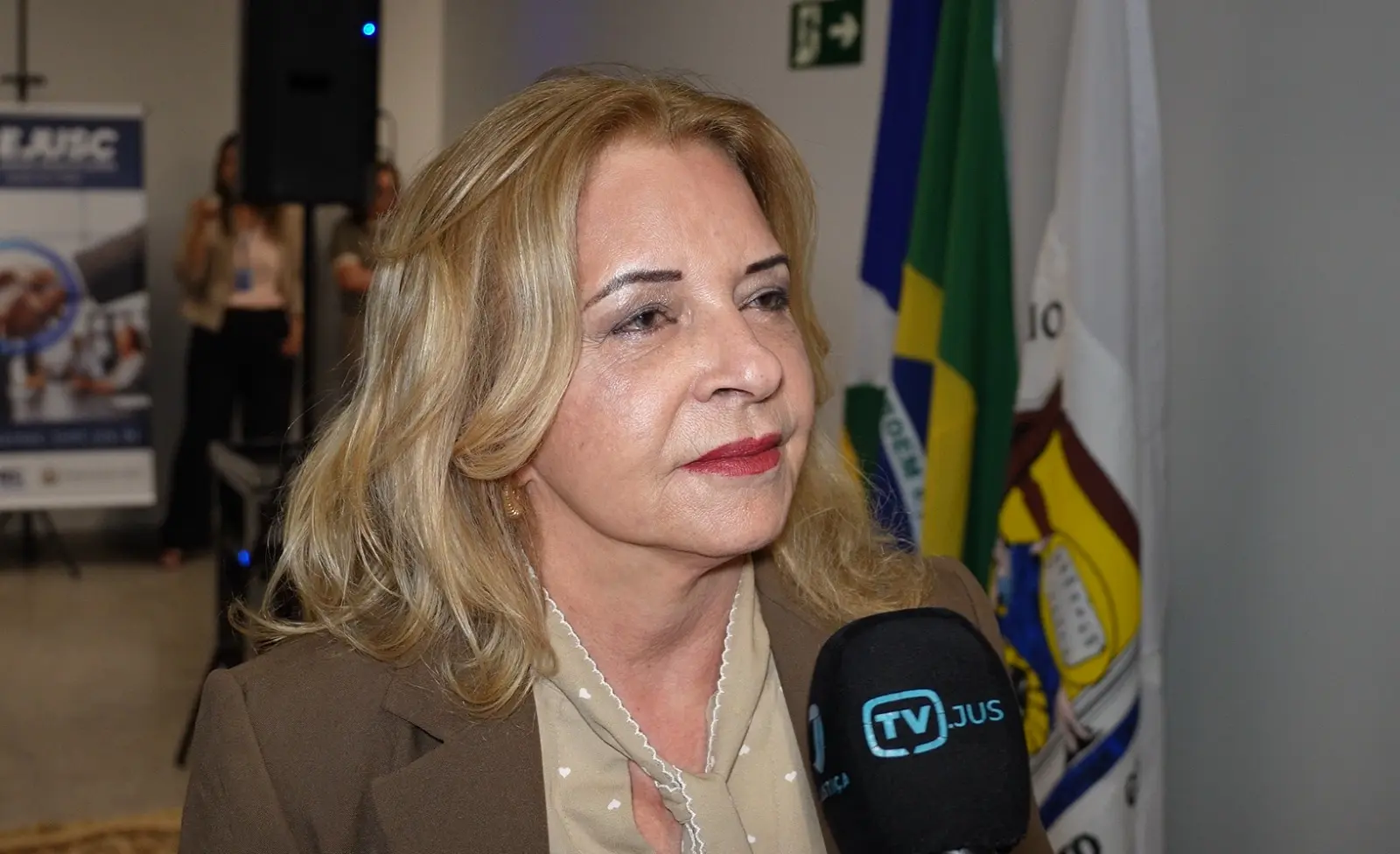 Foto horizontal que mostra a procuradora de justiça Eliana Maranhão do busto pra cima. Ela é uma senhora branca, de cabelos loiros, lisos, na altura dos ombros, usando camisa e blazer em tons sépia. Ela está no saguão do Complexo dos Juizados.