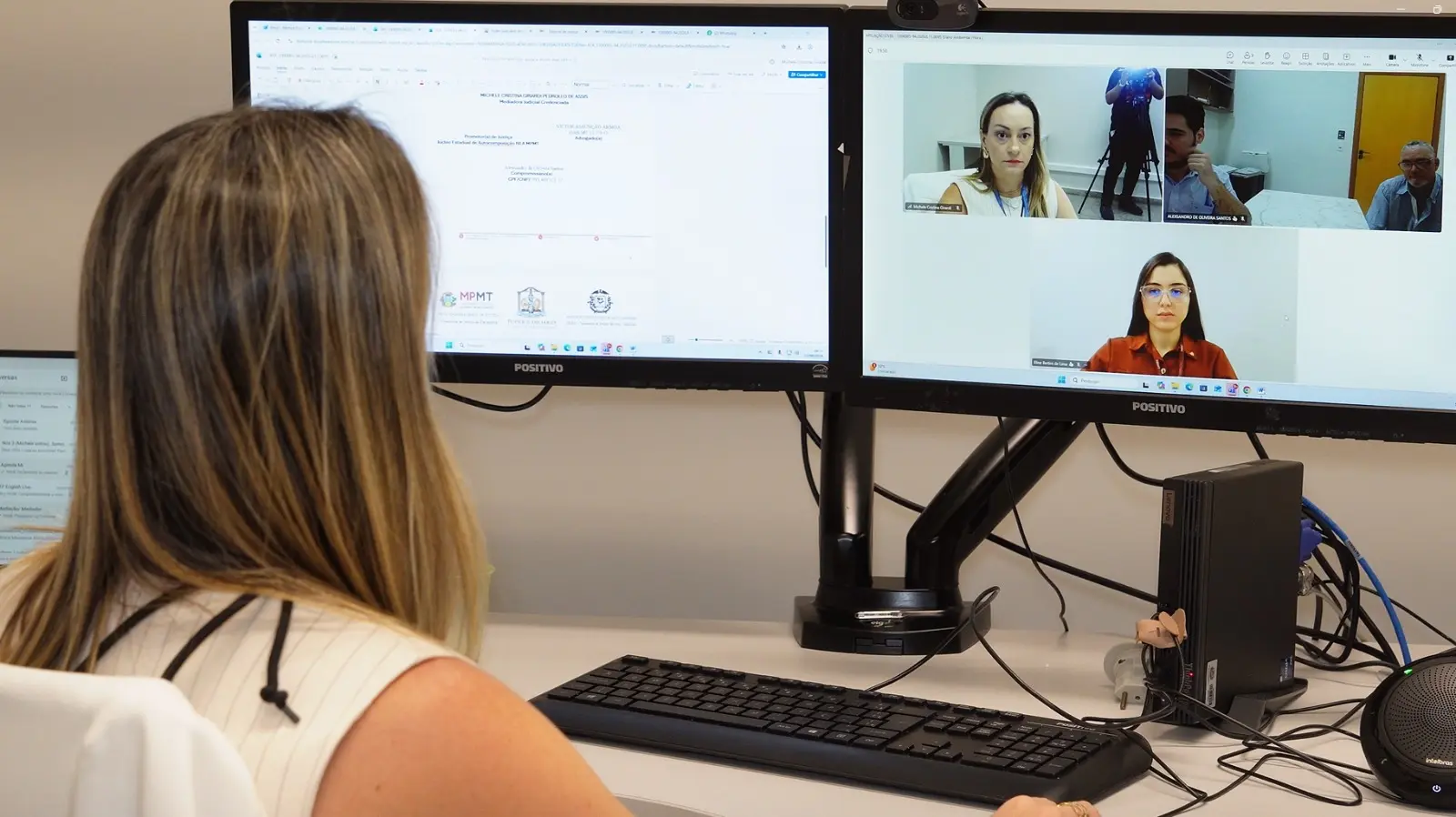 Foto horizontal que mostra uma mulher de costa, em frente ao computador, participando de audiência de conciliação on-line. Na tela, aparecem os participantes da audiência.