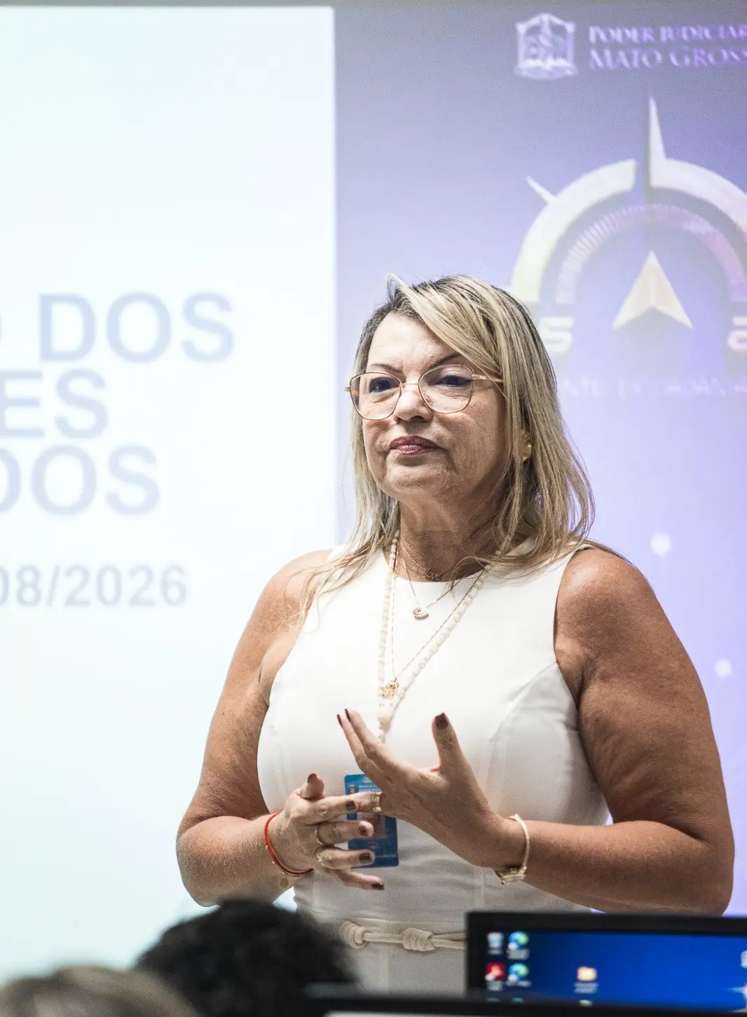 Foto vertical que mostra a gestora da Contadoria do Fórum de Cuiabá, Angélica Vilalva Guimarães, e instrutora da capacitação, fala aos participantes durante a aula. Em pé, ela gesticula enquanto apresenta conteúdos da formação, com projeção institucional ao fundo.