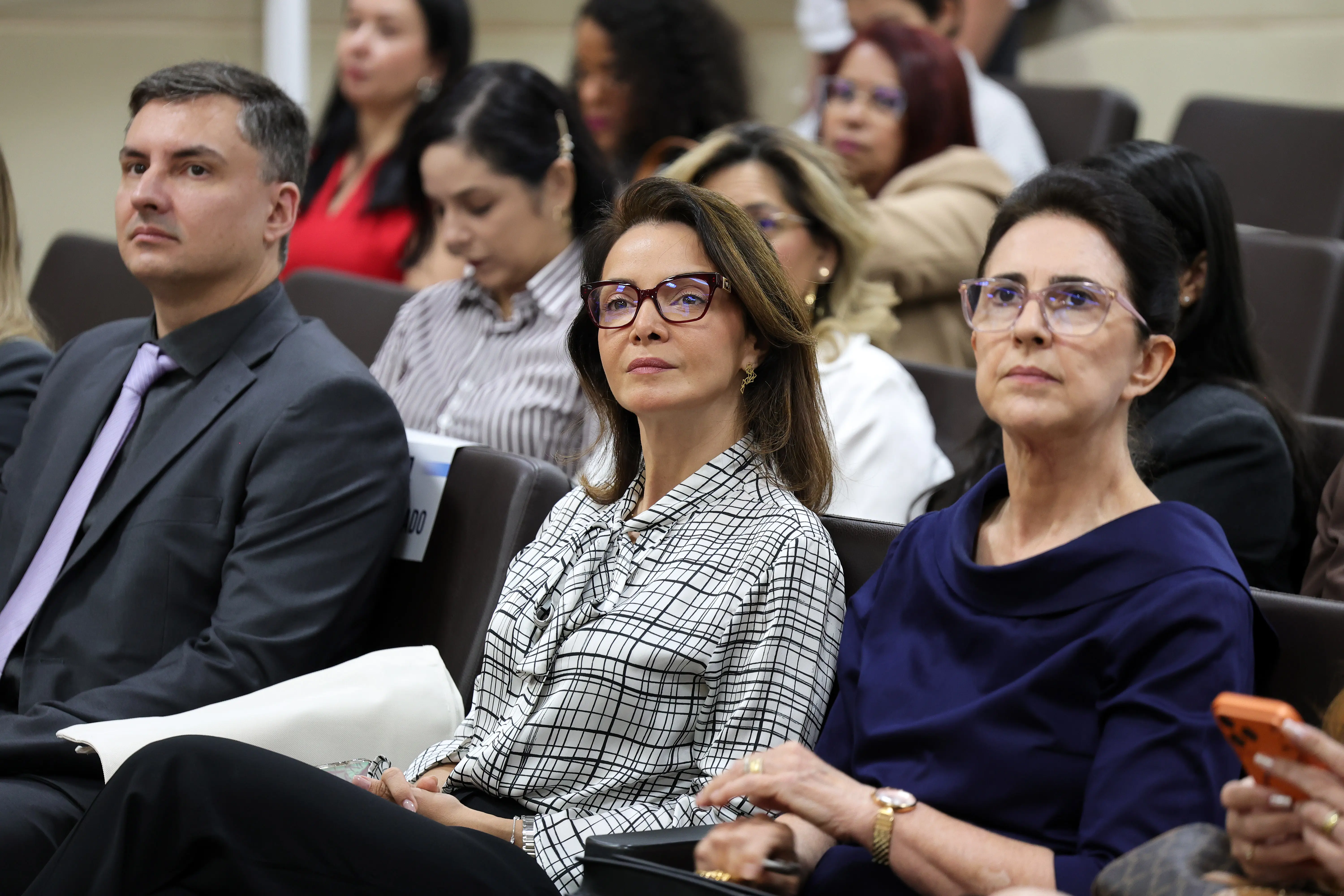 Três pessoas sentadas na plateia de um auditório olham para a frente. À esquerda, homem de terno cinza; ao centro, mulher de óculos e blusa xadrez; à direita, mulher de óculos e vestido azul-escuro.