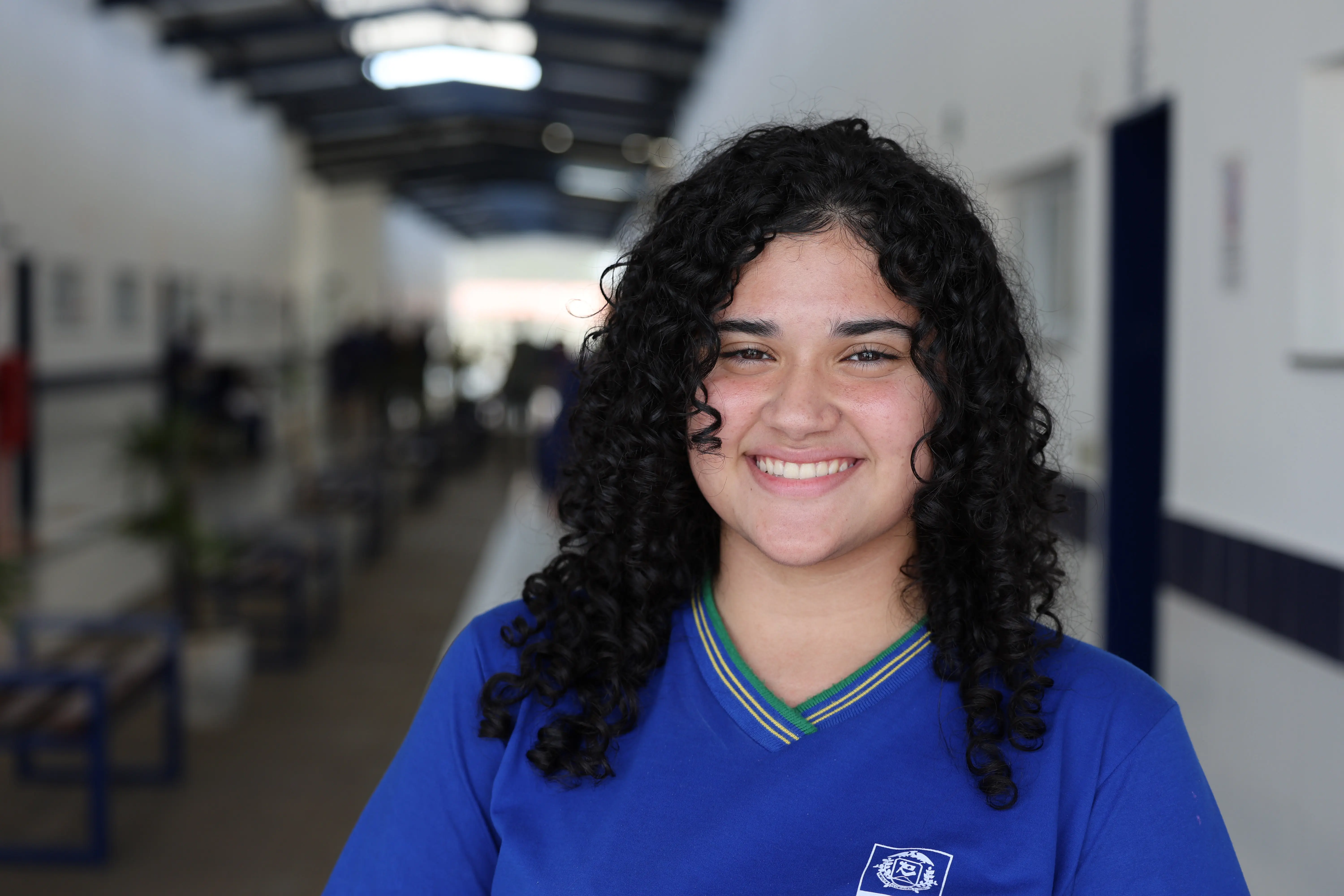 Retrato de Ana Cristina Olímpio. Jovem de cabelos pretos, longos e bem cacheados. Ela sorri abertamente para a câmera, vestindo a camiseta azul do uniforme estadual. O corredor da escola aparece desfocado ao fundo.