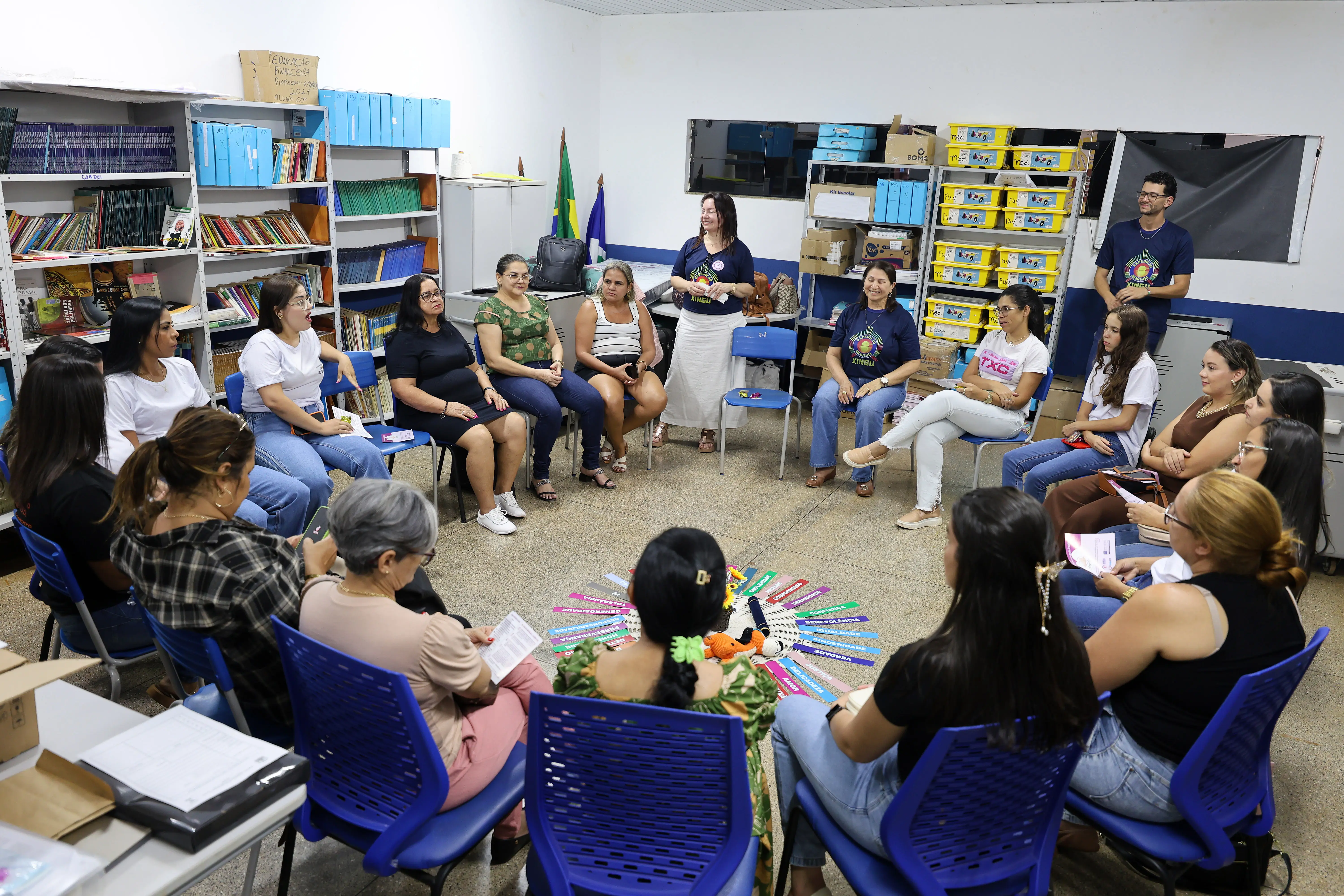 Pessoa gesticulando em sala organizada; prateleiras com caixas amarelas etiquetadas. Espaço funcional, voltado para ações comunitárias ou educativas.