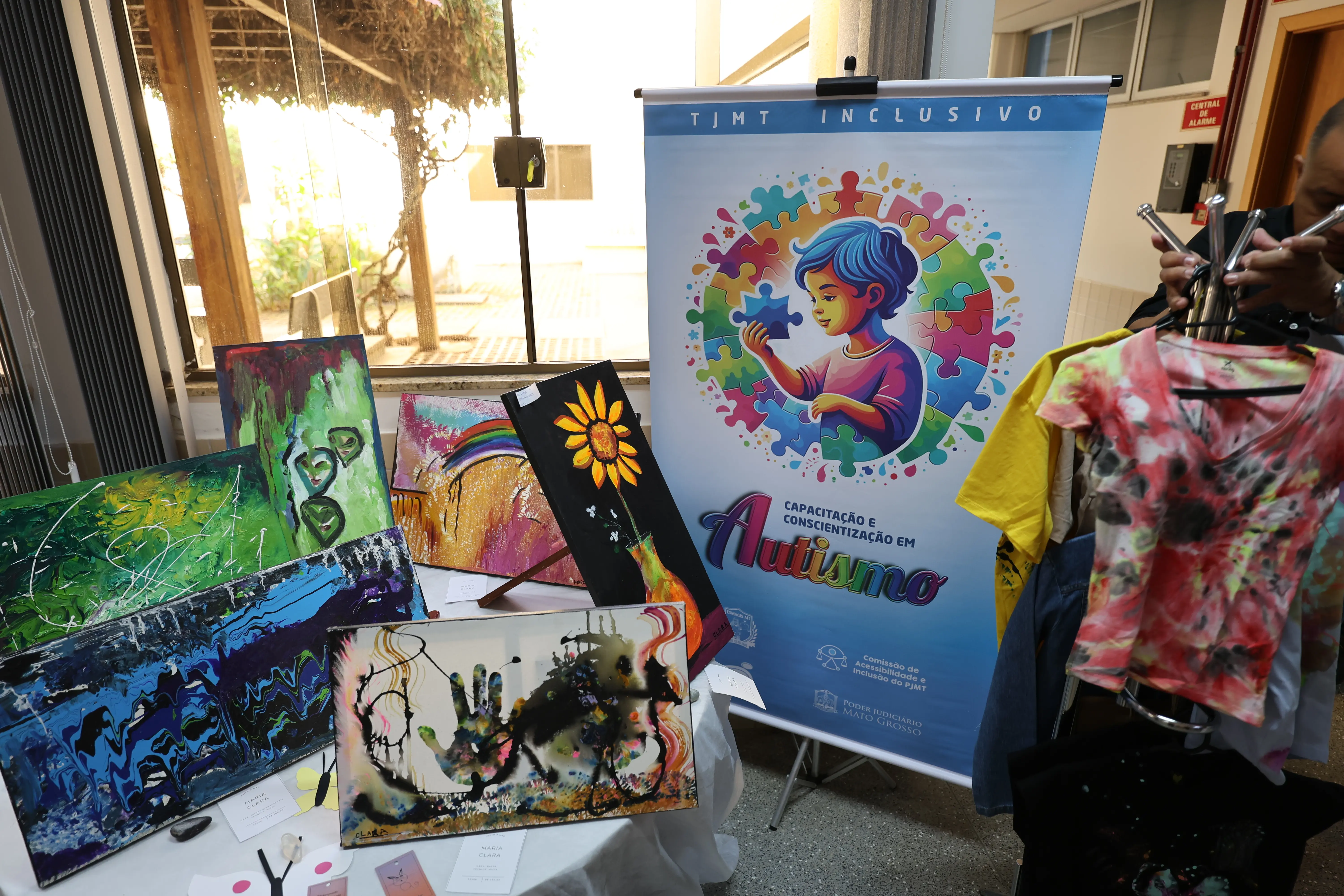 Painel do TJMT Inclusivo destaca o tema autismo ao lado de coloridas obras de arte e camisetas pintadas à mão. O espaço iluminado por luz natural valoriza a criatividade e a expressão artística dos participantes.