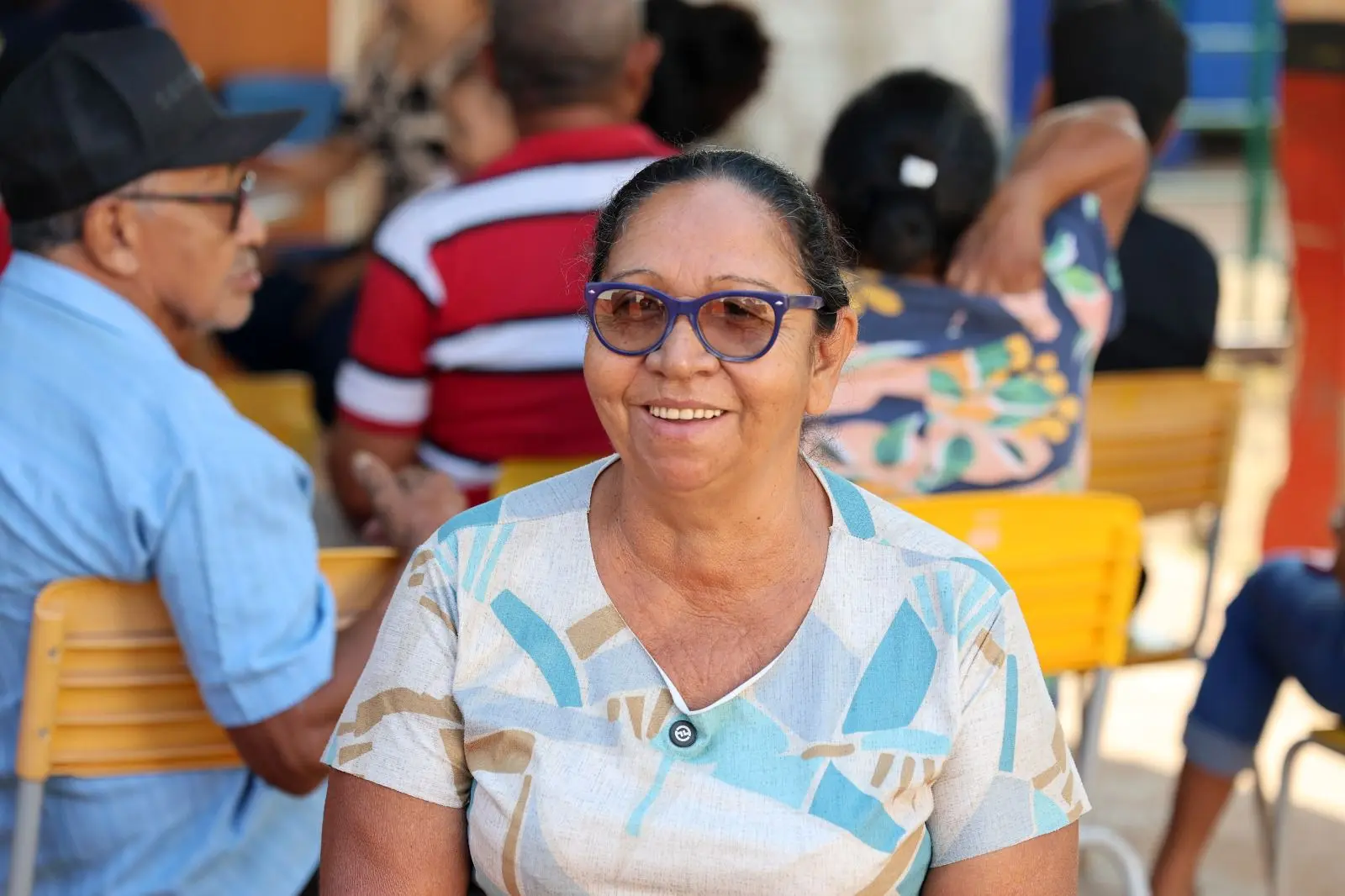 Mulher idosa de óculos azuis e vestido estampado em tons claros sorri sentada em cadeira amarela. Ao fundo, outras pessoas aguardam atendimento em espaço simples e iluminado, com clima comunitário e acolhedor.